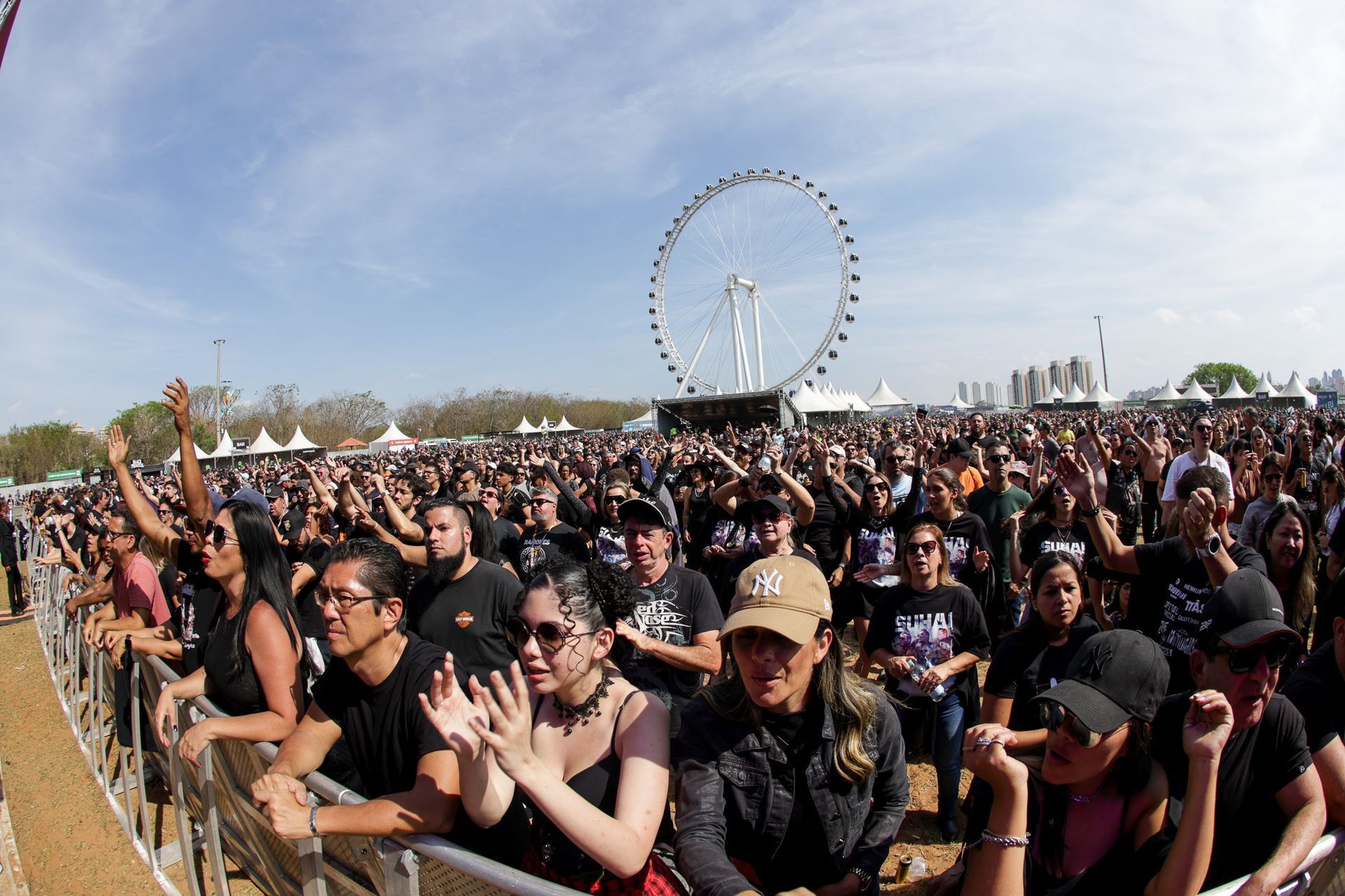 Uma multidão se reúne em frente a uma grande roda-gigante em um festival de música ao ar livre.