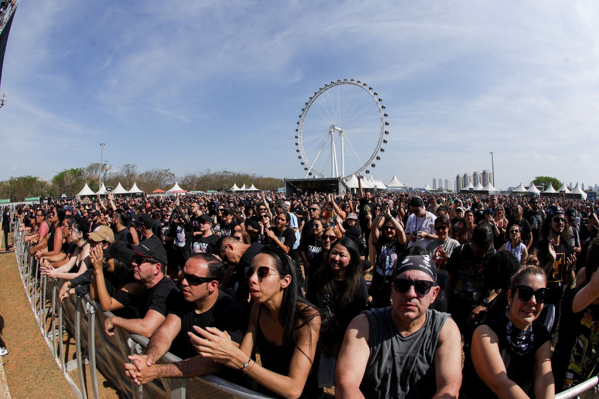 Uma grande multidão em um festival de música está em pé diante de uma barreira, com uma roda-gigante gigante ao longe.