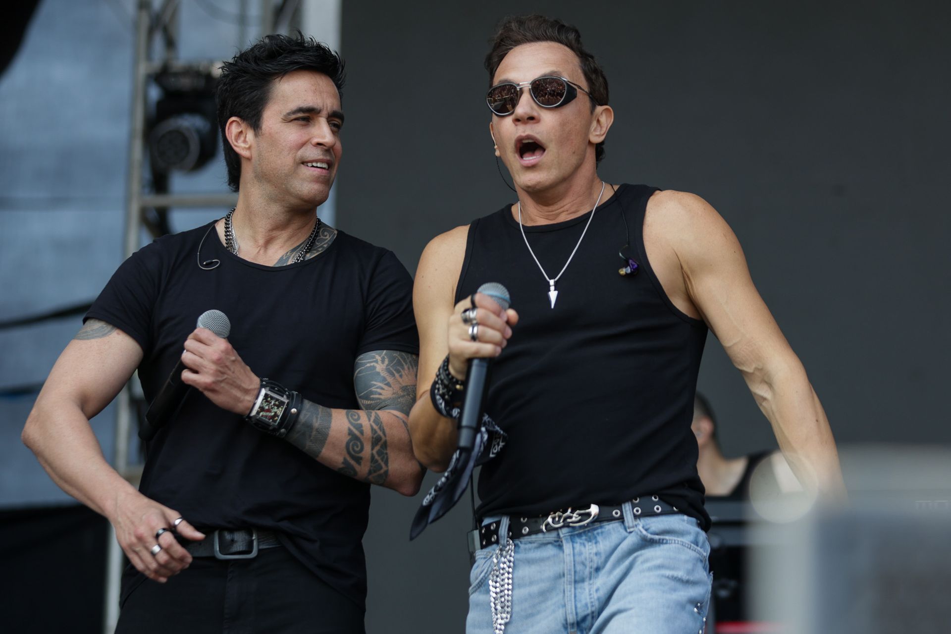 Dois homens se apresentando no palco. Ambos vestem camisetas pretas sem mangas, colares e pulseiras, e estão cantando.