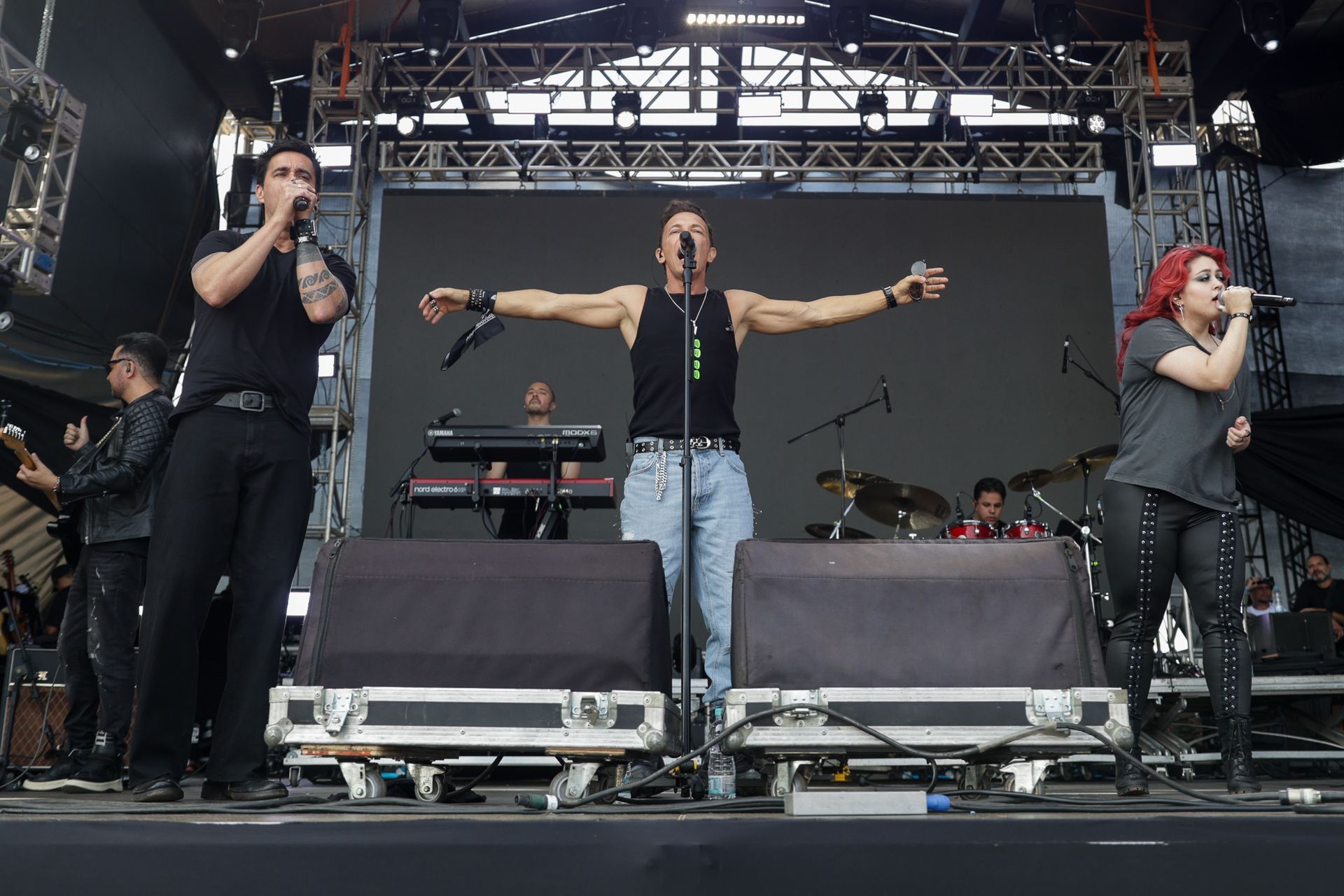 Uma banda se apresenta em um palco ao ar livre, com o vocalista estendendo os braços enquanto os outros cantam em microfones.