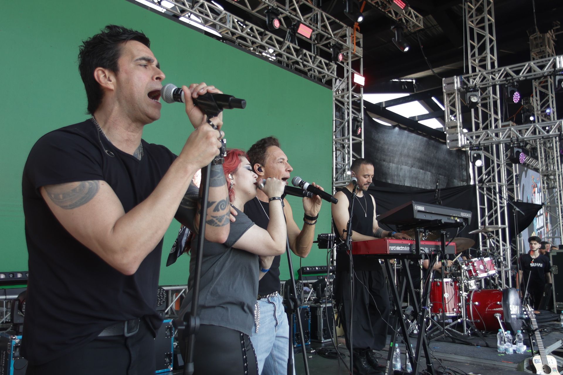 Uma banda se apresenta em um palco ao ar livre; os vocalistas cantam em microfones enquanto um tecladista toca.