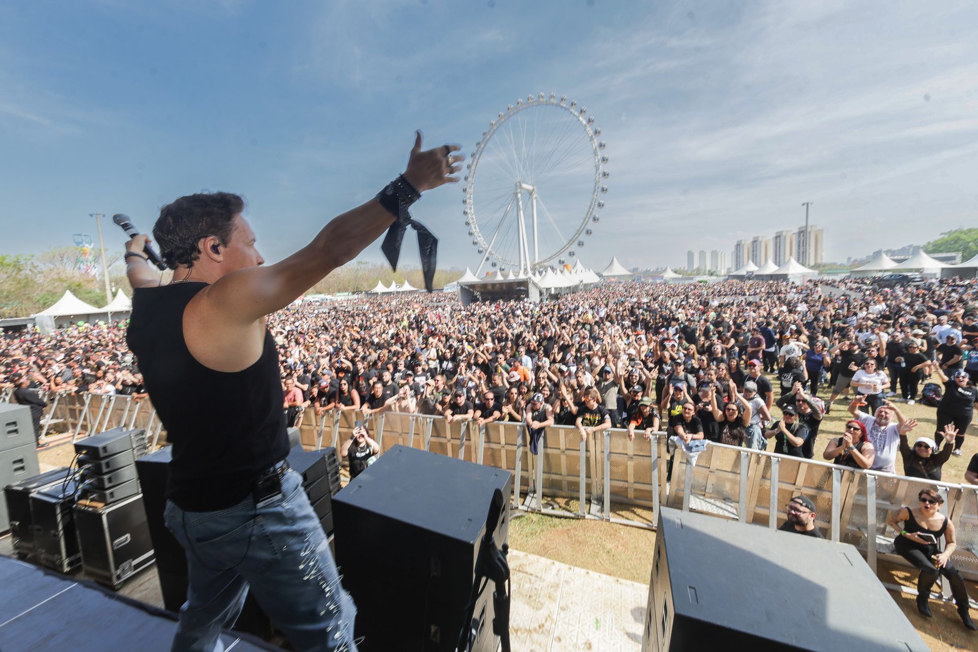 Um artista no palco, de frente para uma multidão grande em um festival ao ar livre, com uma roda-gigante ao fundo.
