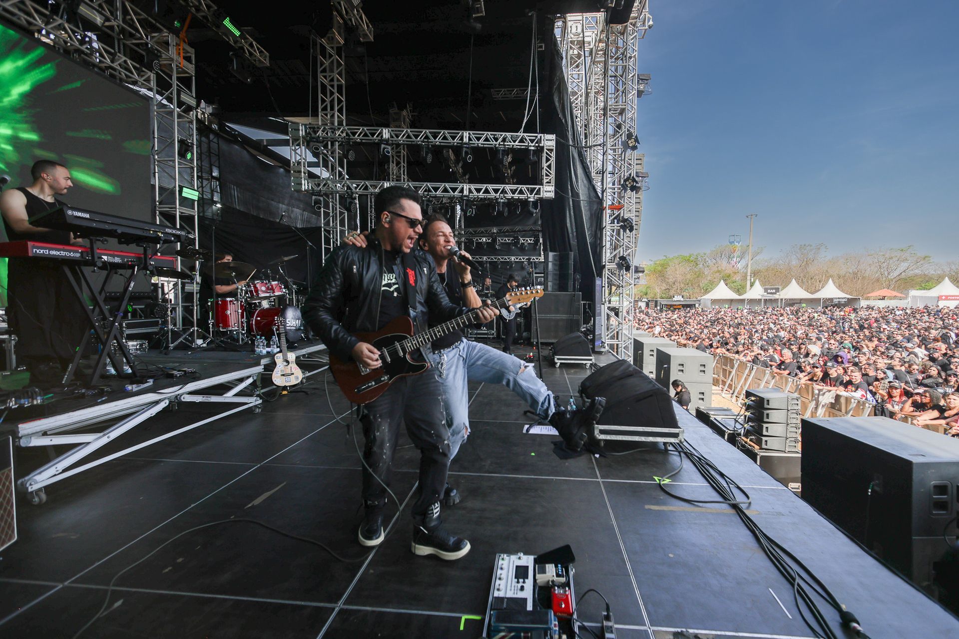 Uma banda se apresenta em um palco para uma grande multidão em um festival de música ao ar livre em um dia ensolarado.