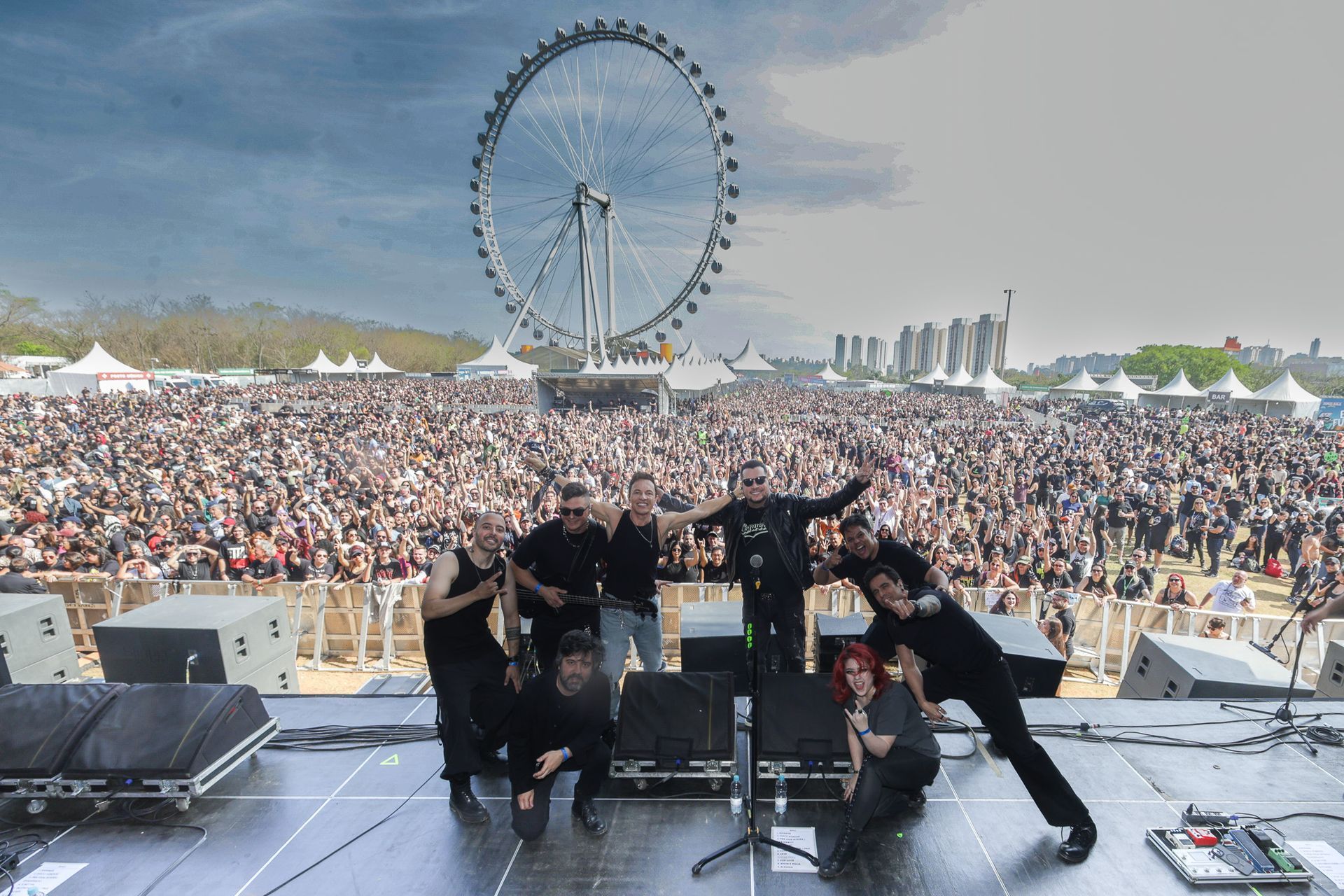Uma banda posa em um palco diante de uma grande multidão animada em um festival, com uma roda-gigante gigante ao fundo.