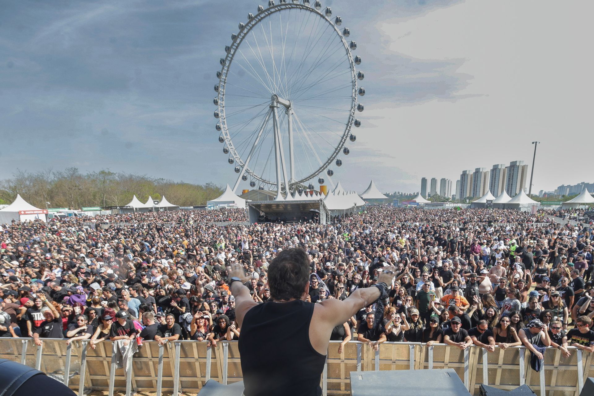 Um artista no palco com os braços erguidos, de frente para uma multidão em um festival, tendo como fundo uma roda-gigante.