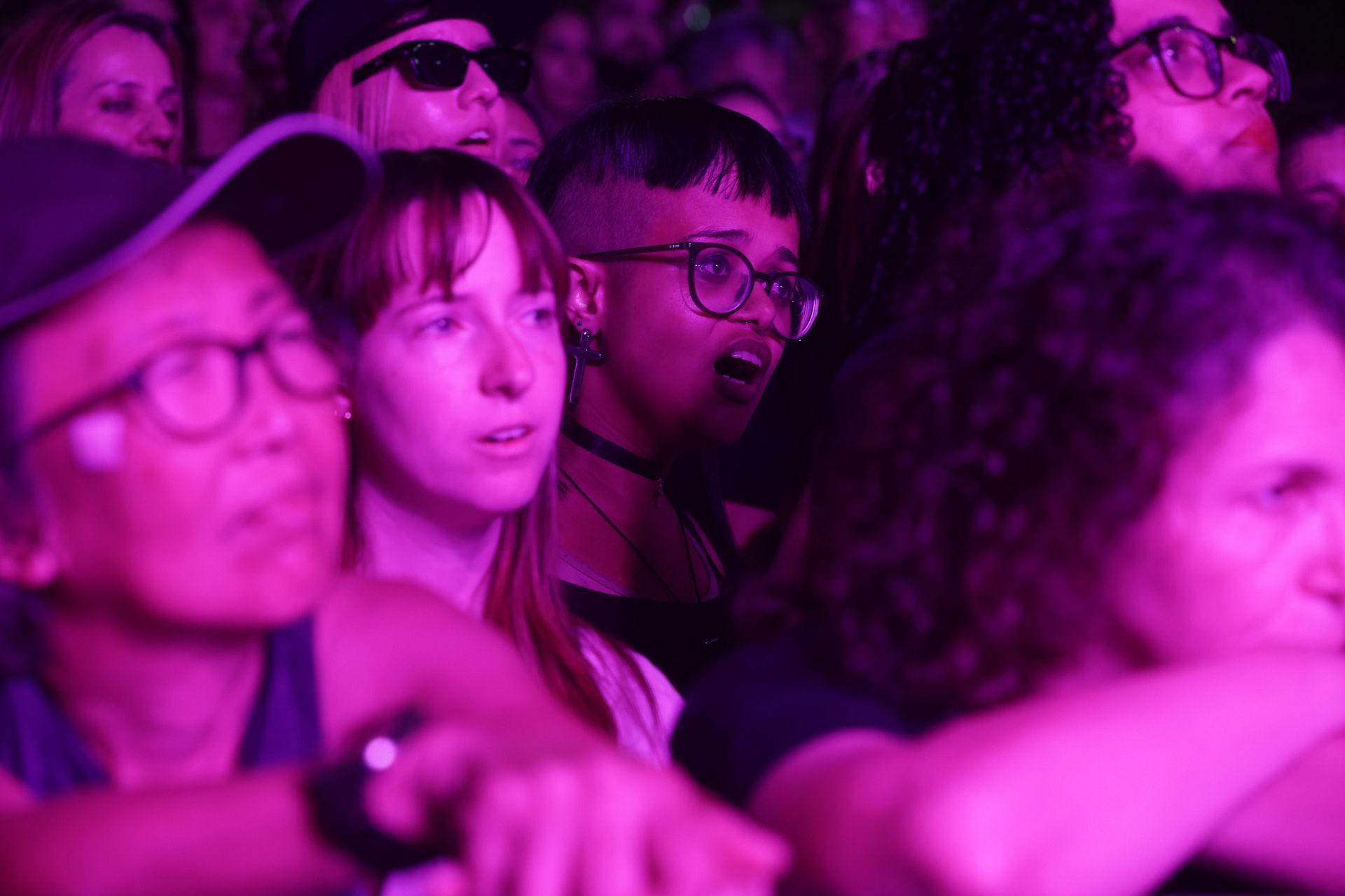 Pessoas em uma multidão, iluminadas por luz rosa, assistindo a uma apresentação. Os rostos mostram expressões de entusiasmo