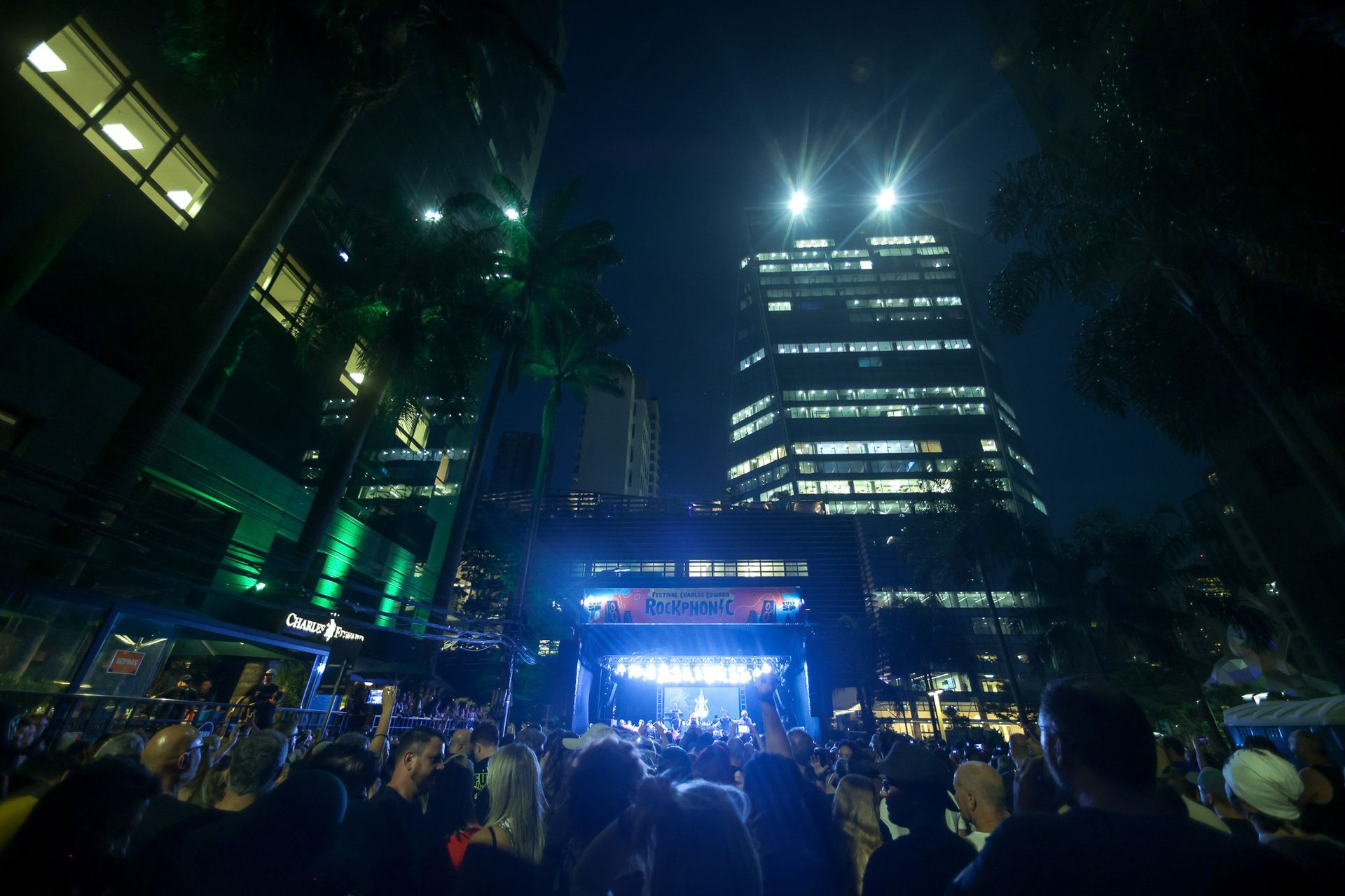 Concerto noturno ao ar livre em uma praça da cidade com palco iluminado e público, tendo como pano de fundo prédios altos.
