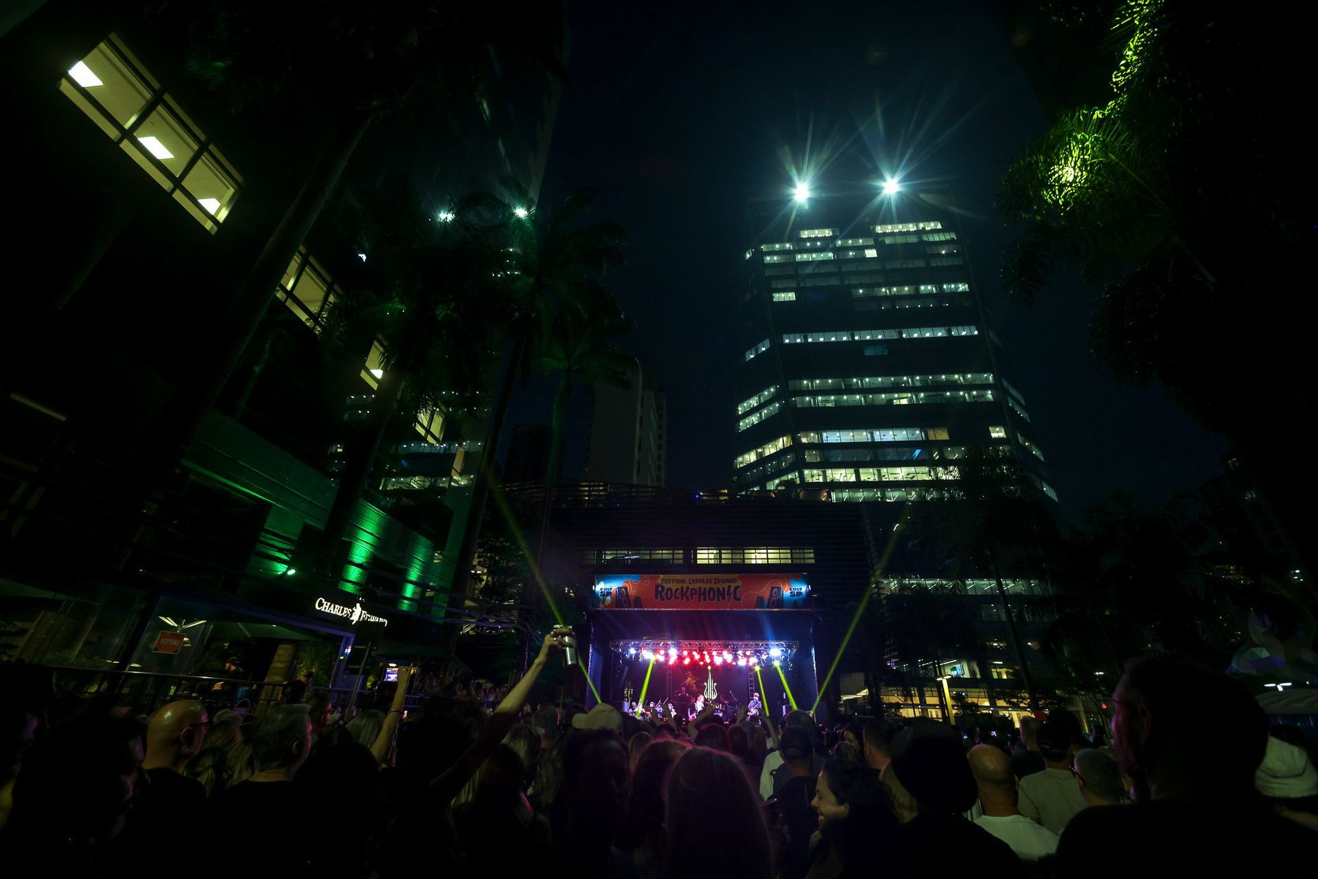 Concerto noturno ao ar livre, palco com luzes, público, prédios da cidade.