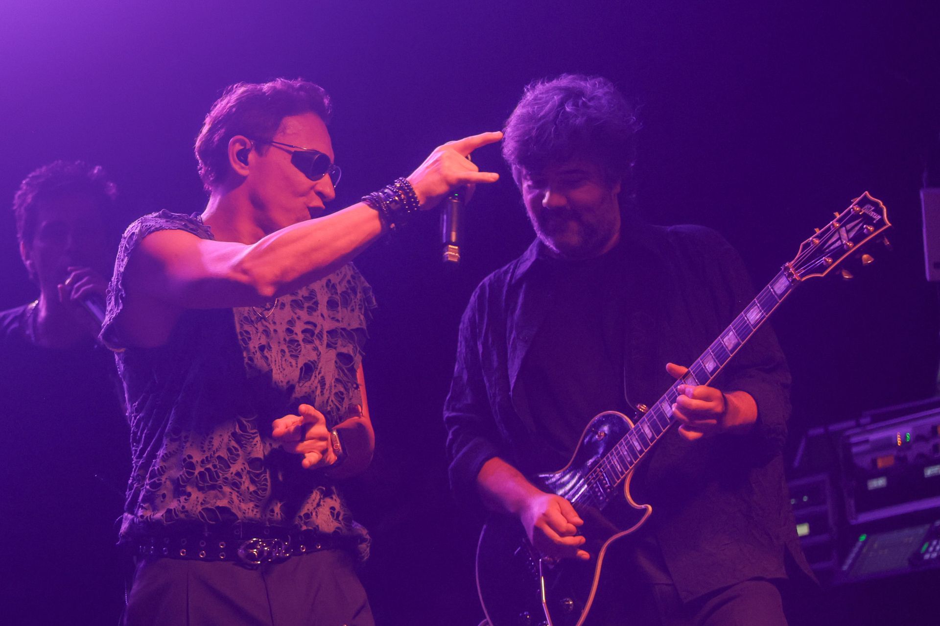Cantor de óculos escuros aponta para a cabeça do guitarrista, ambos no palco sob luz roxa. O guitarrista toca.