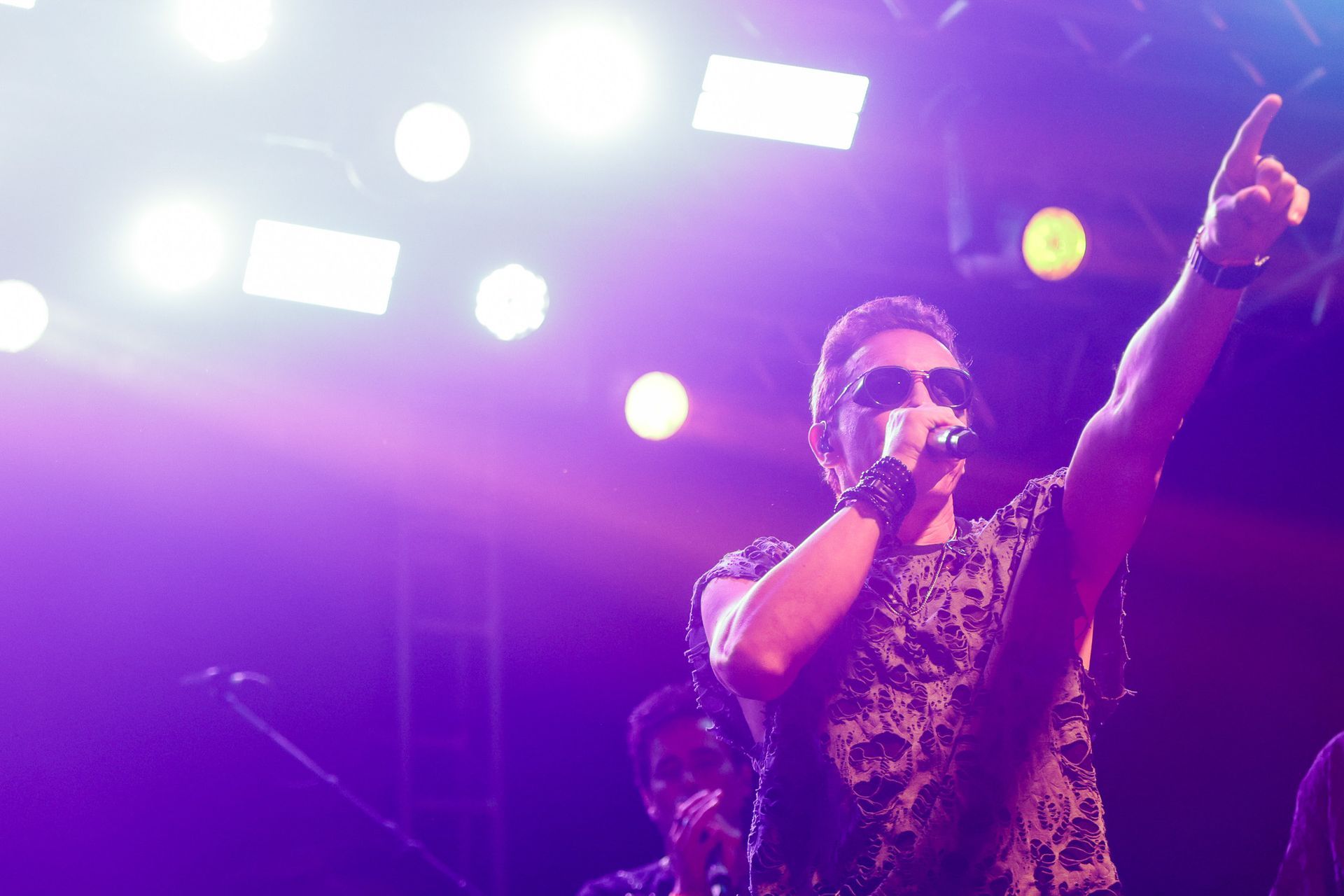 Um cantor de óculos escuros e microfone no palco, apontando para cima. Luzes de palco roxas.