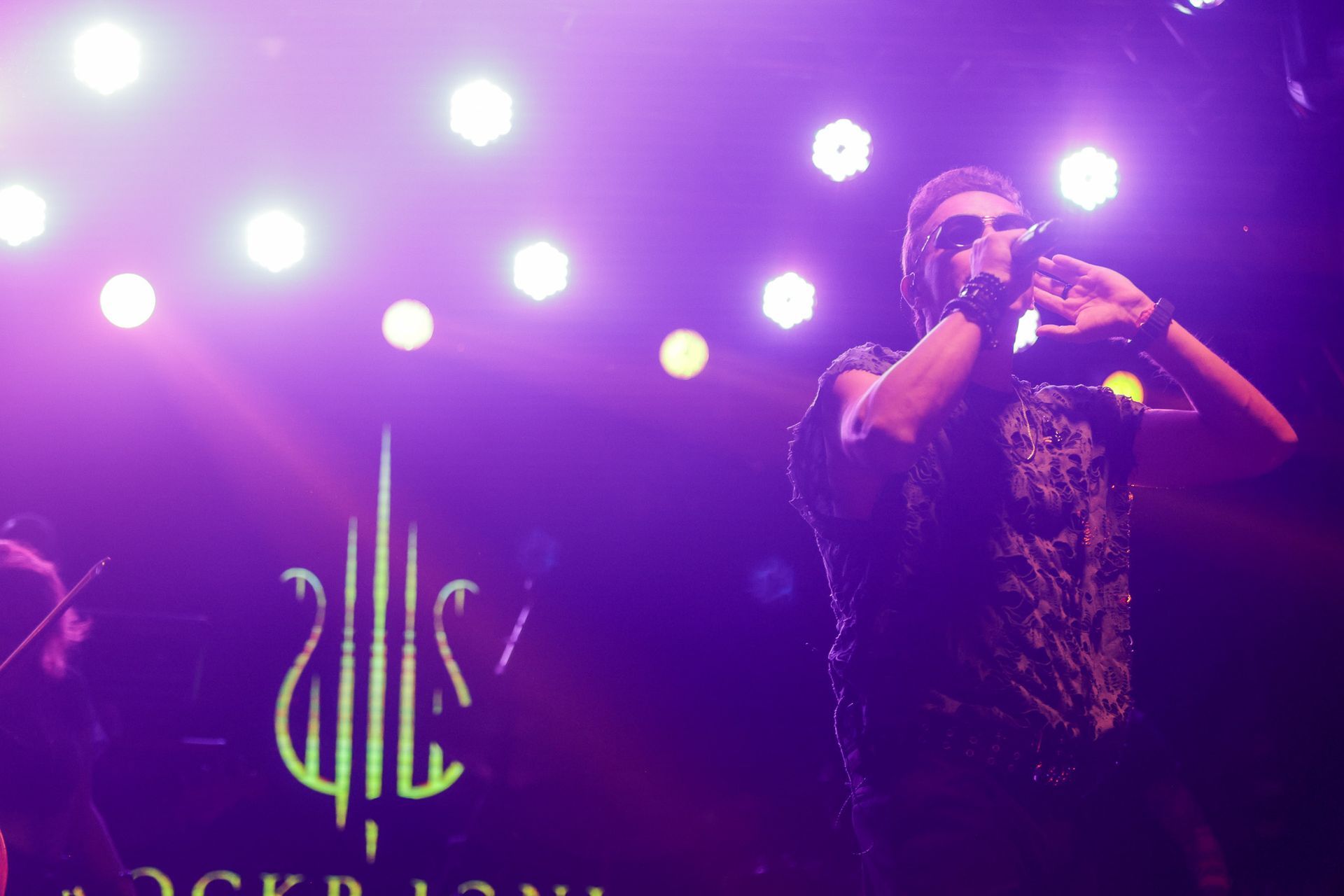 Cantor no palco com microfone, iluminação de palco roxa, logotipo da banda ao fundo.