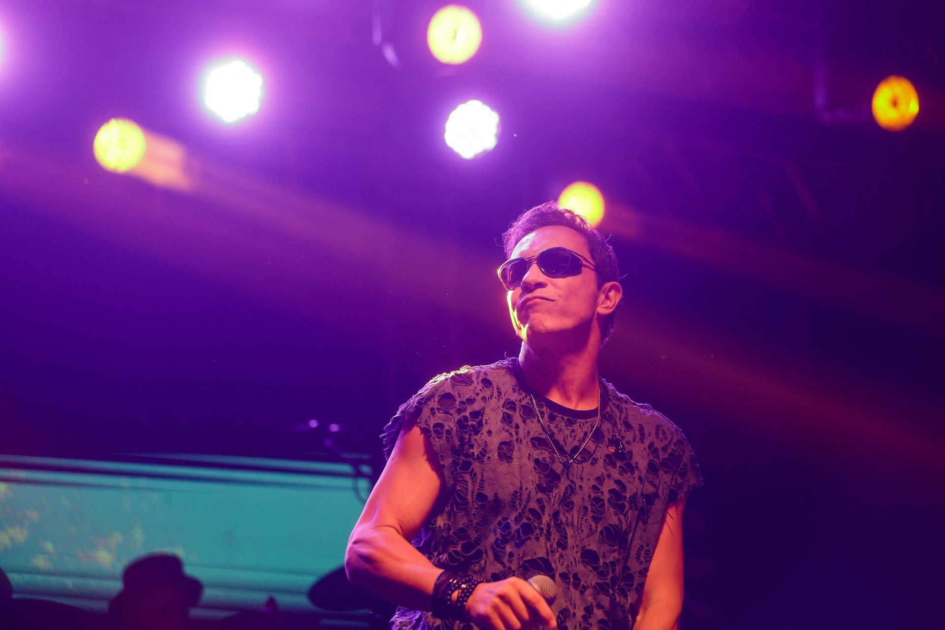 Um artista de óculos escuros no palco, cantando ao microfone sob as luzes do palco.