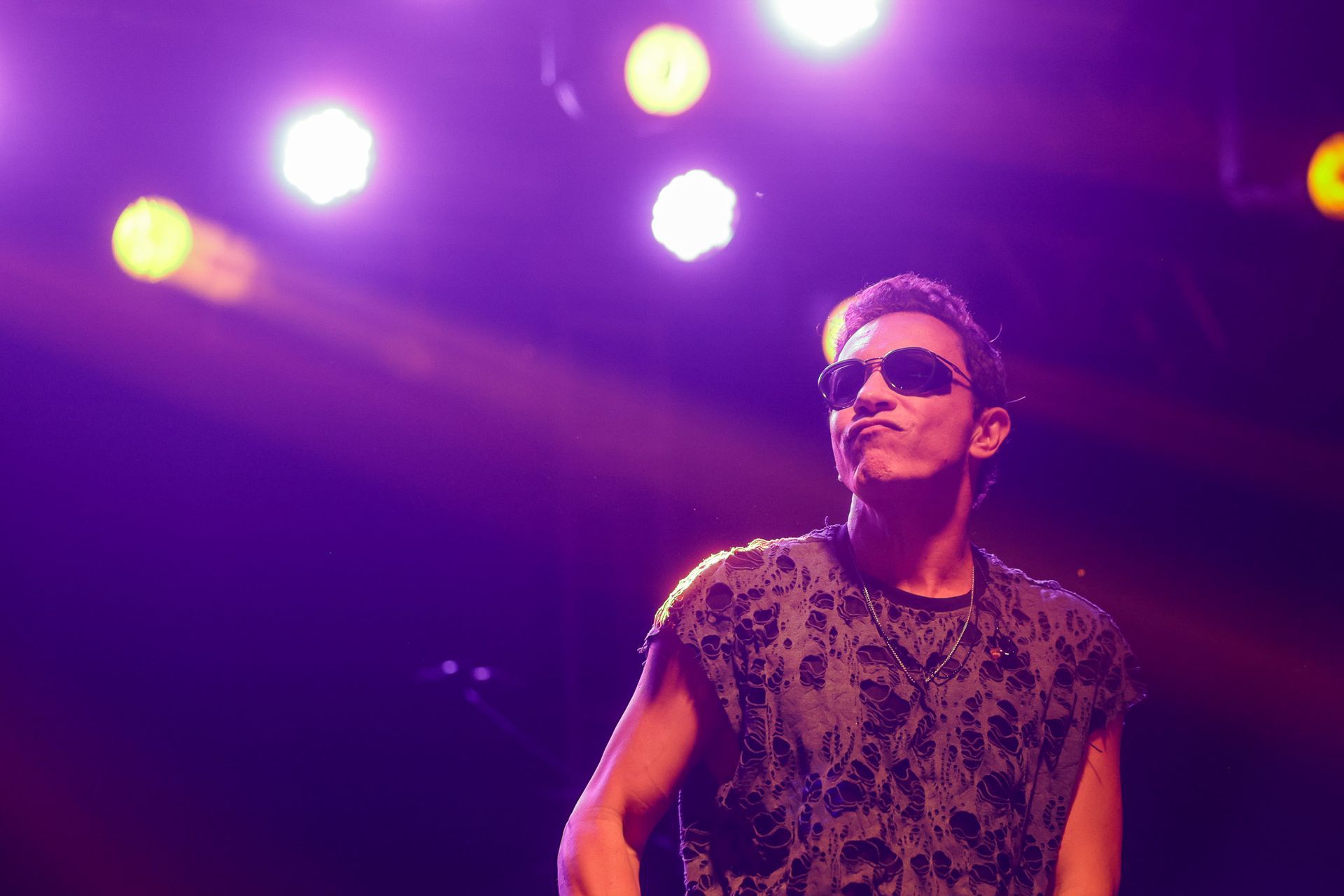 Homem de óculos escuros, no palco com luzes roxas, olhando para cima.