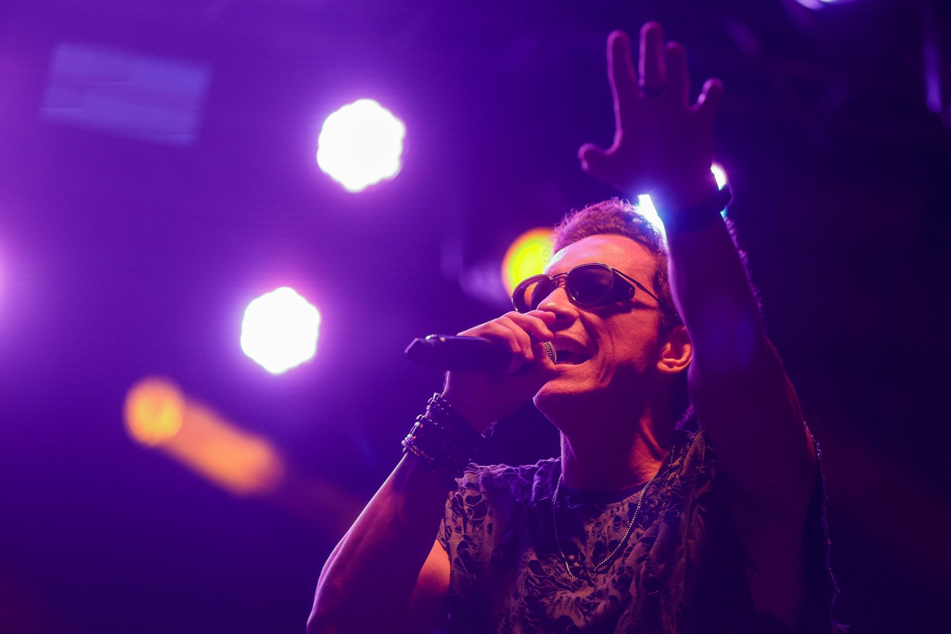 Cantor no palco com microfone, braços erguidos, sob holofotes roxos.