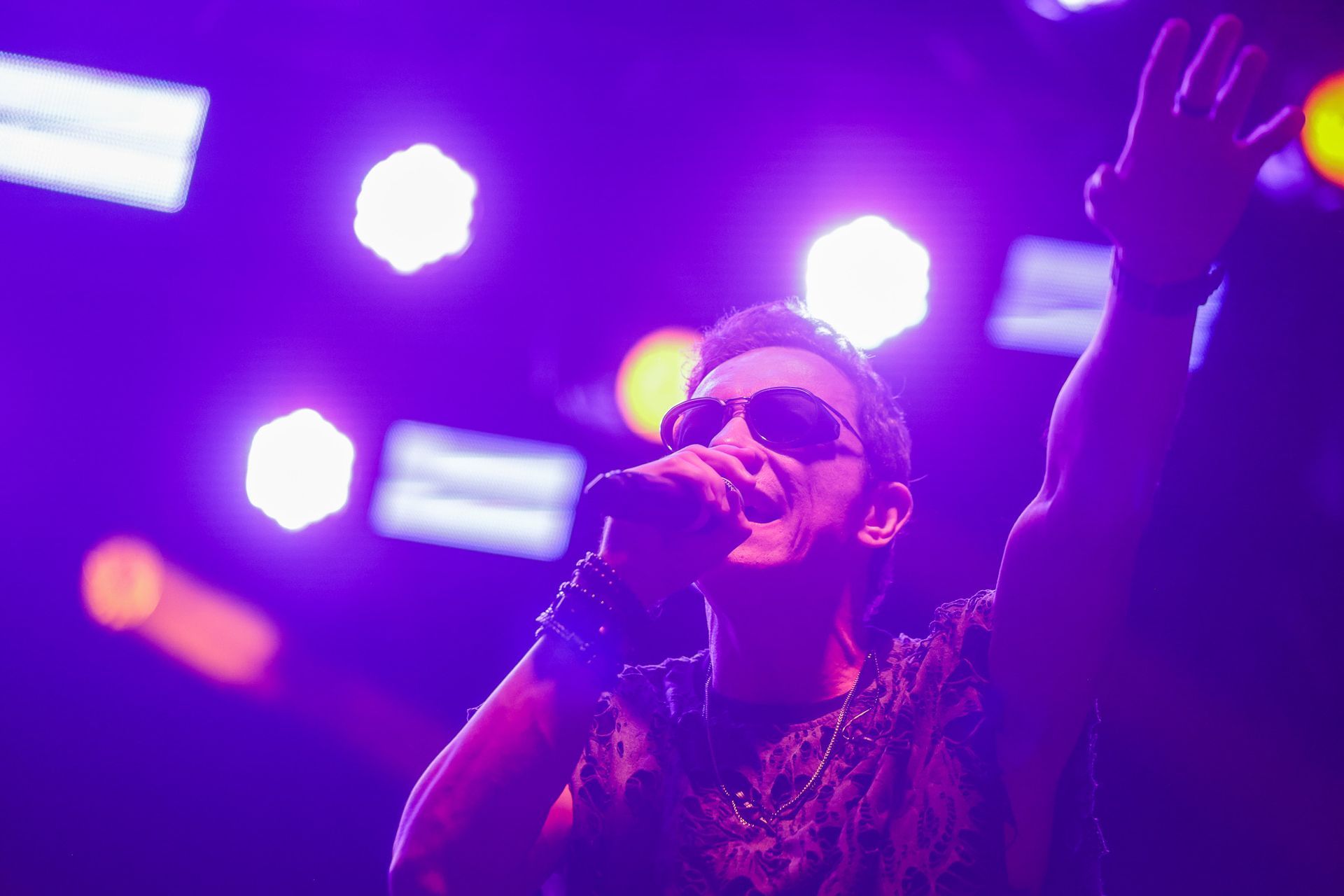 Cantor no palco com microfone, iluminação de palco roxa, logotipo da banda ao fundo.