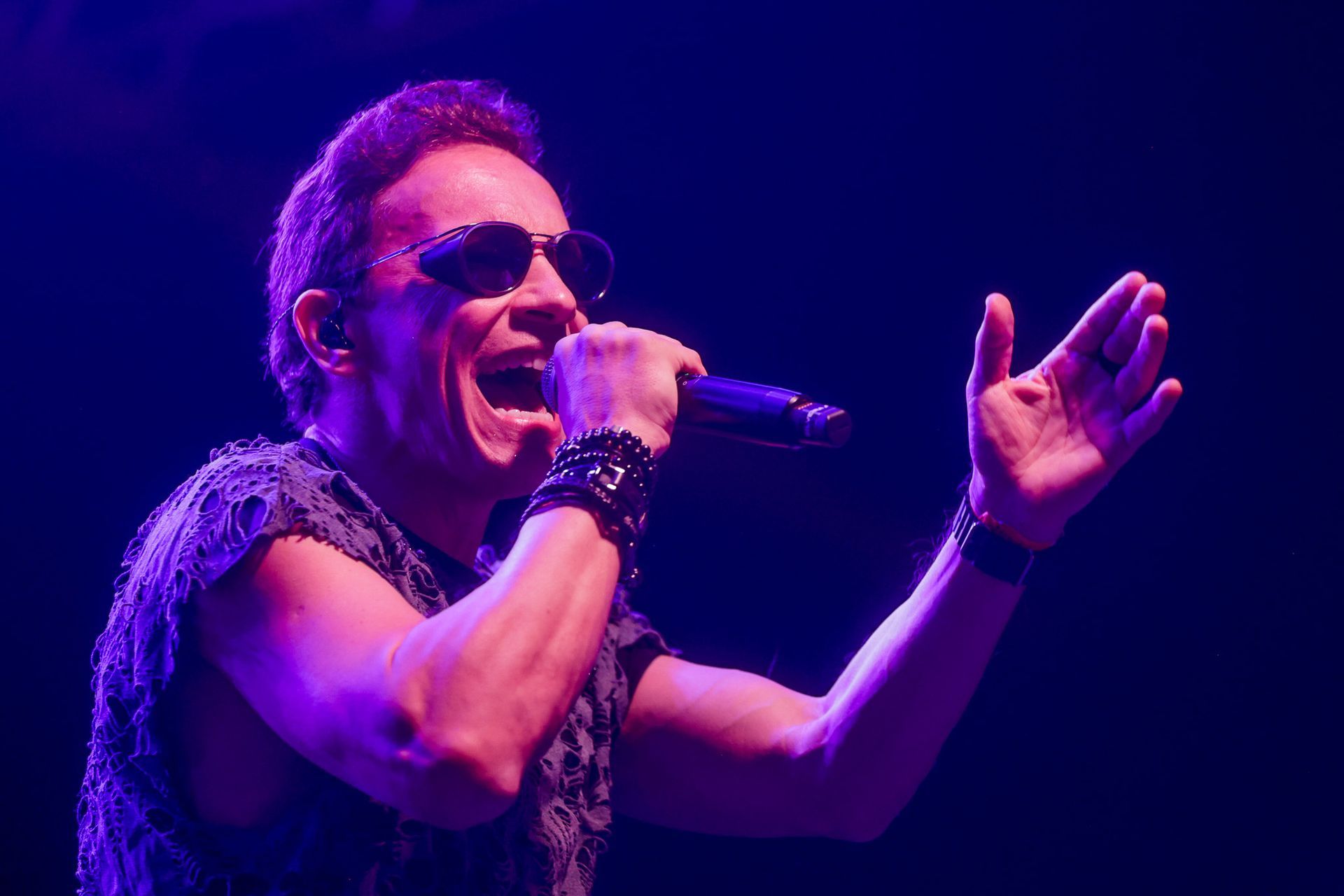 Cantor de óculos escuros e microfone, se apresentando no palco com o braço levantado