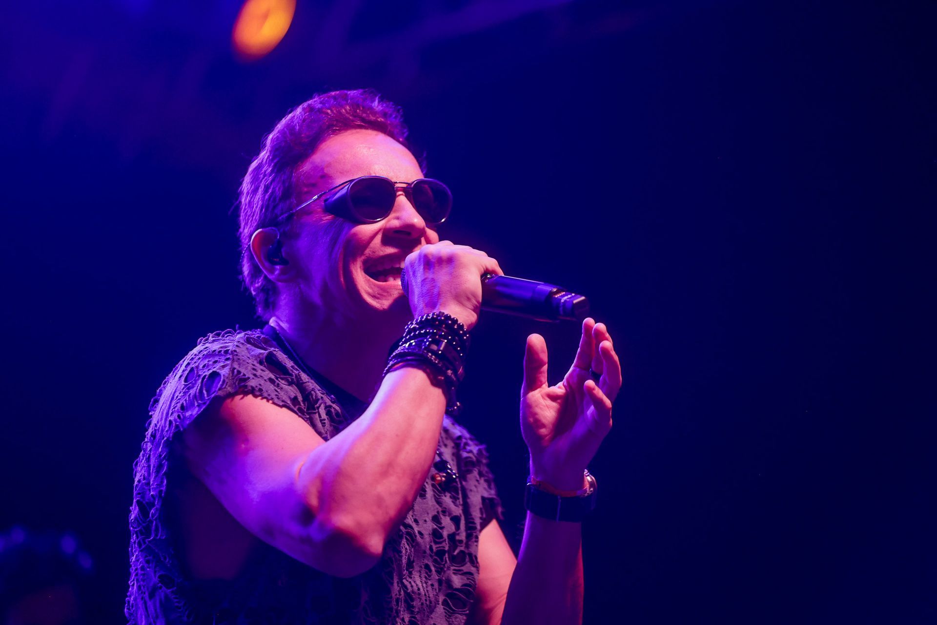 Homem cantando em um microfone no palco, usando óculos escuros, camiseta preta sem mangas e pulseiras; 