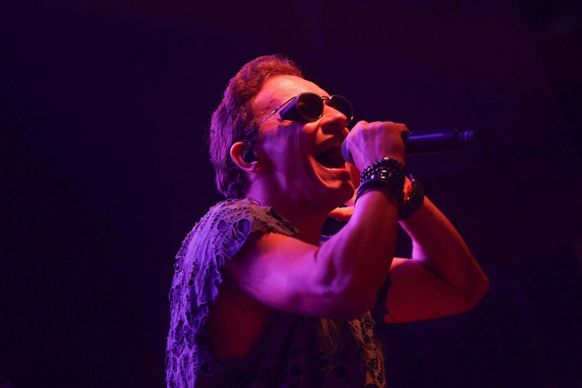 Cantor de óculos escuros, cantando em um microfone, iluminada por luz roxa.