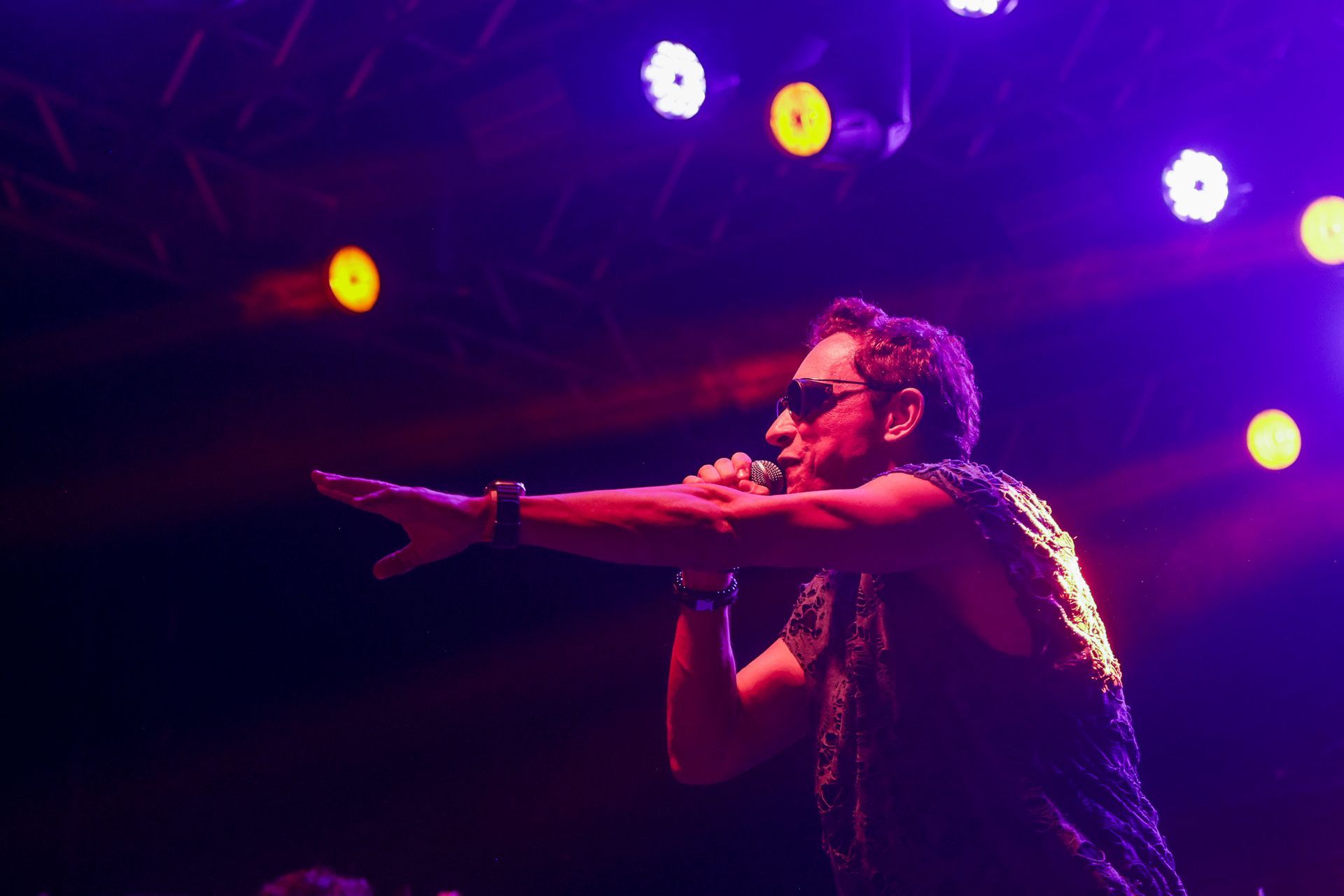 Homem cantando no palco, com o braço estendido. Luzes de palco roxas e amarelas iluminam a cena.