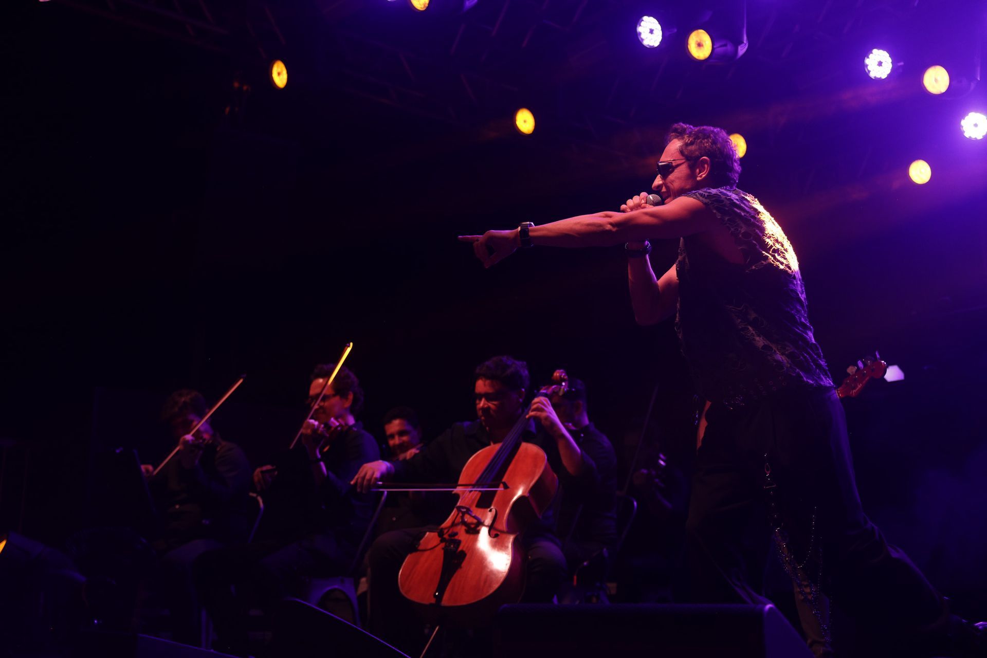 Uma banda se apresenta no palco com uma orquestra, banhada por luzes roxas e brancas.
