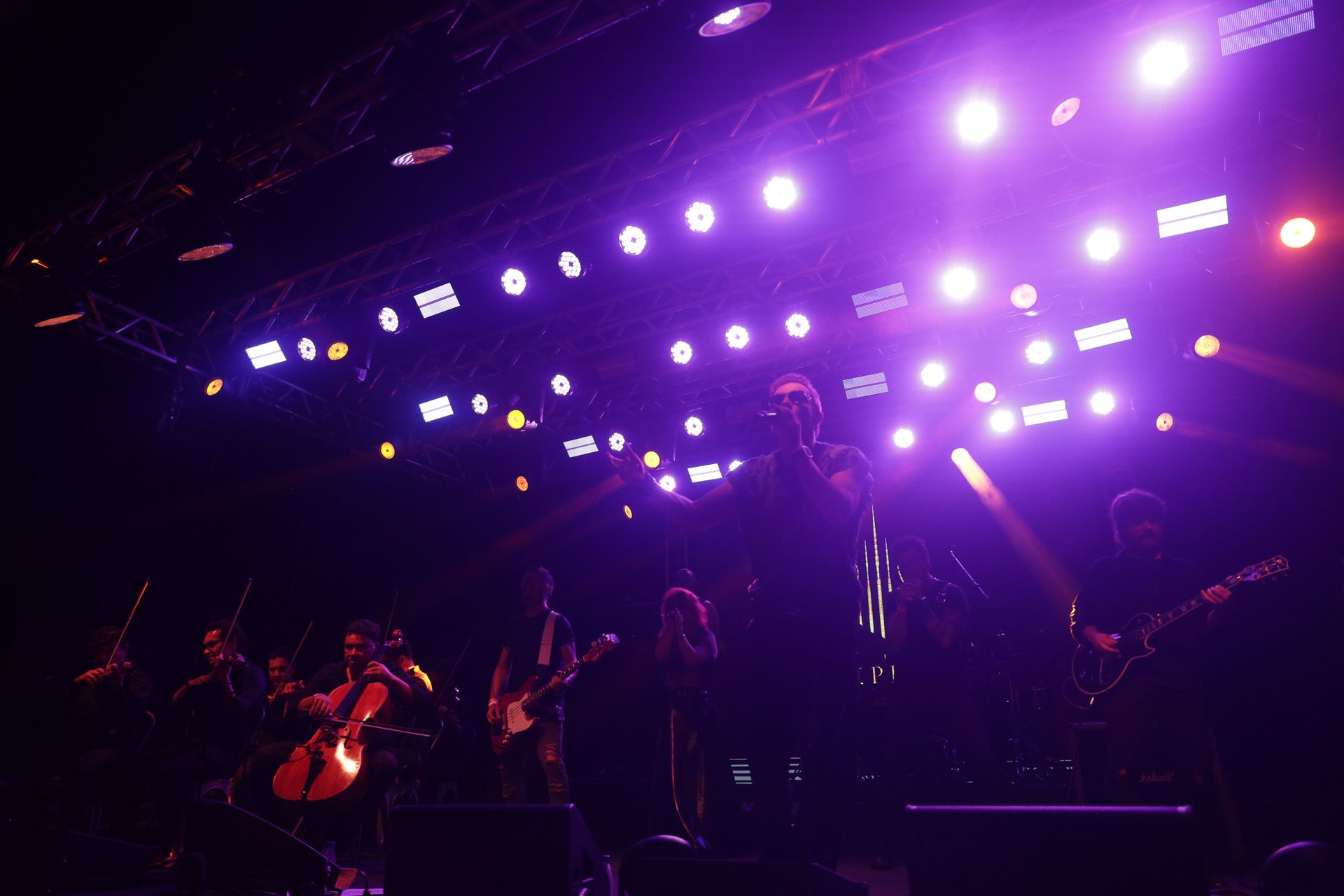 Banda se apresentando no palco, banhada por luzes cênicas roxas e brancas. Músicos tocando instrumentos, em silhueta.
