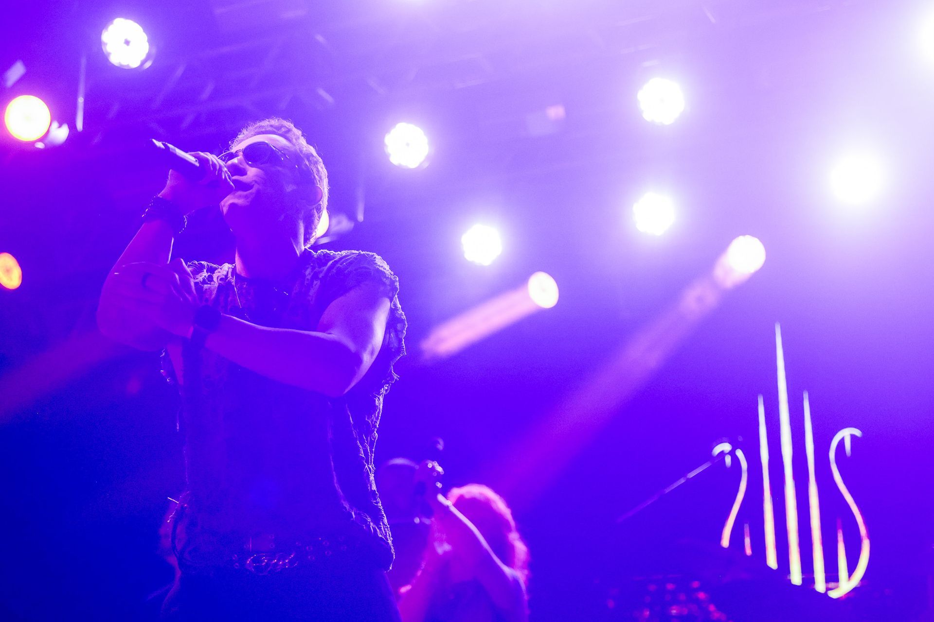 Cantor se apresentando no palco sob luzes de palco roxas e brancas.