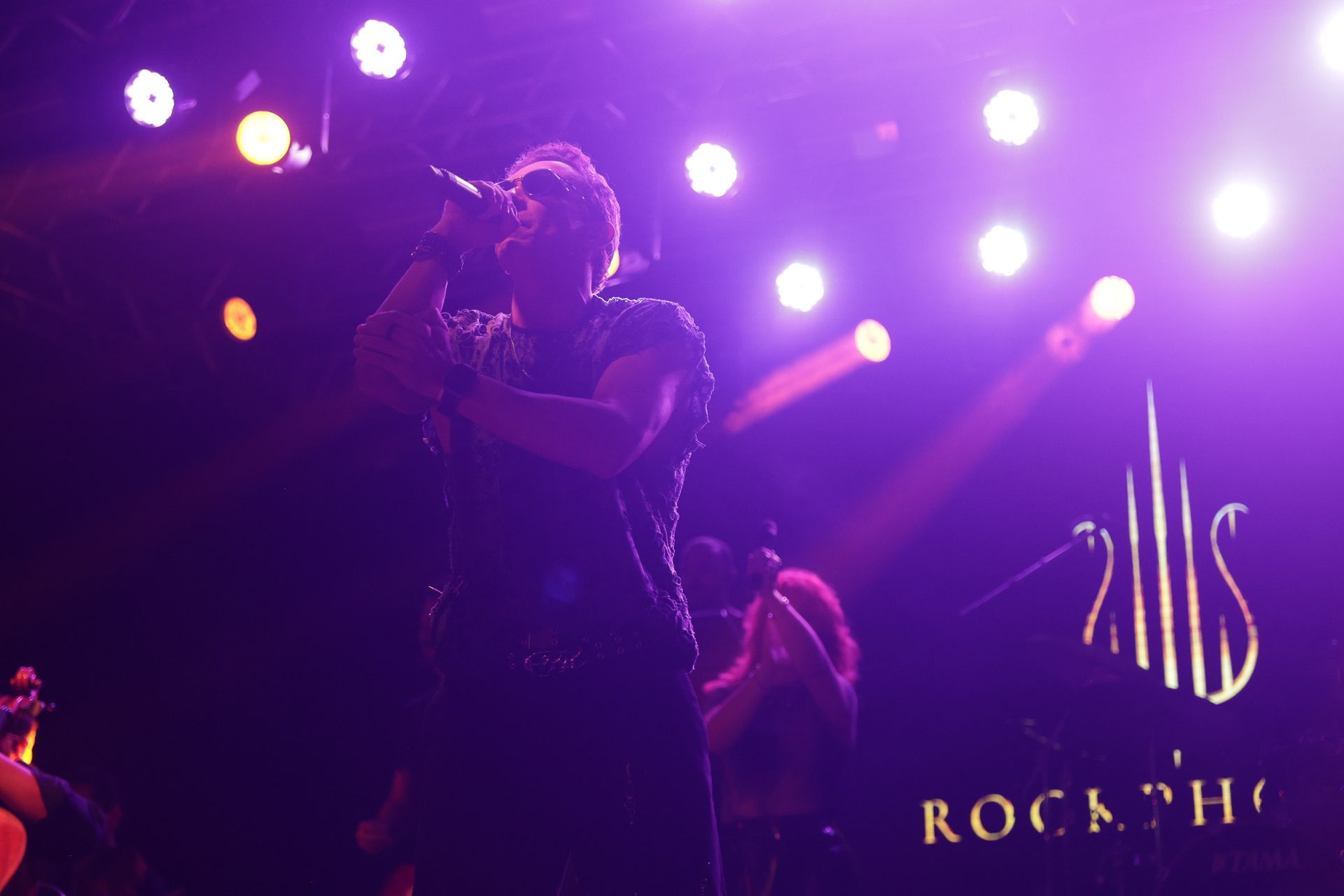 Homem cantando no palco, com o braço estendido. Luzes de palco roxas e amarelas iluminam a cena.