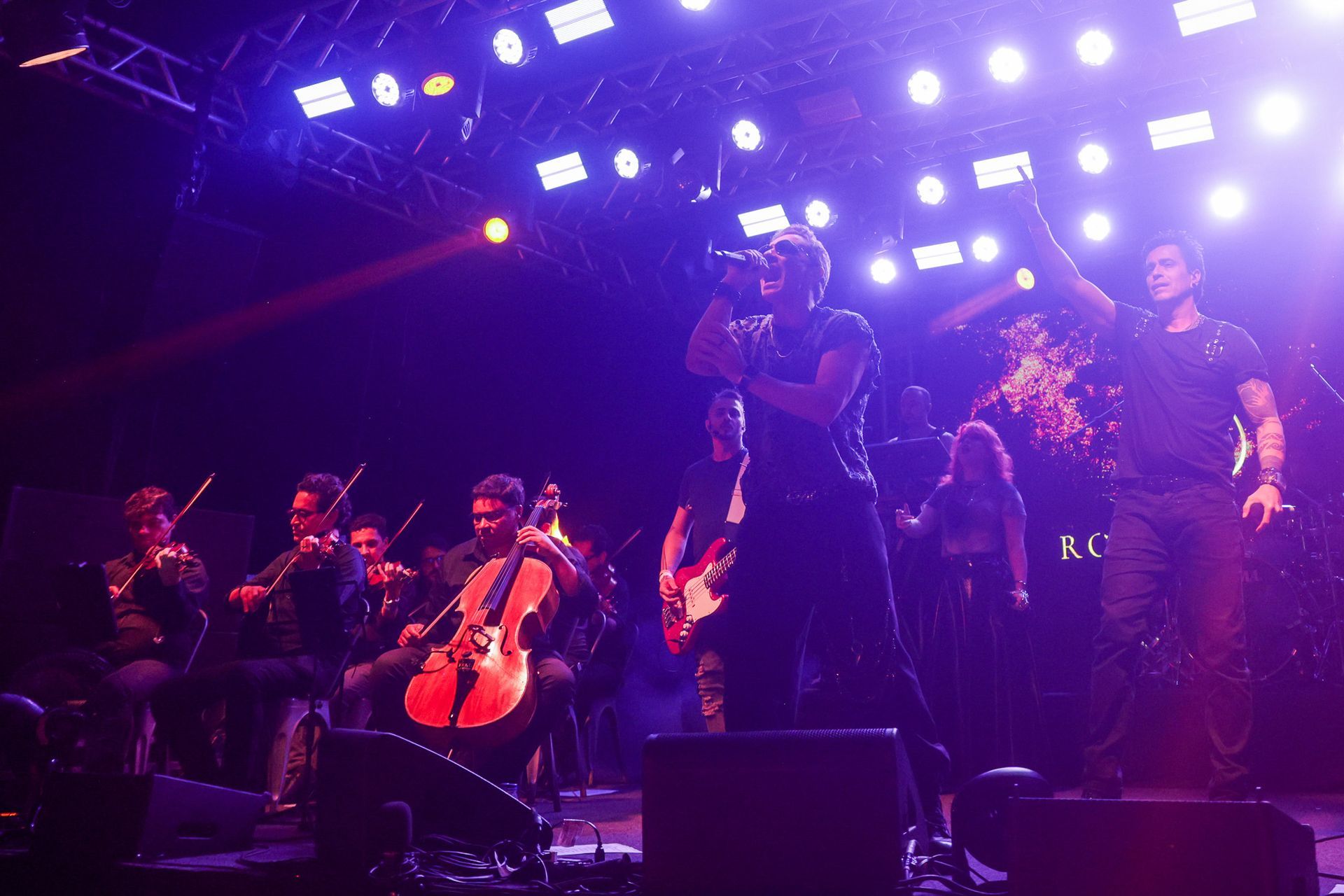 Uma banda se apresenta no palco com uma orquestra, banhada por luzes roxas e brancas.