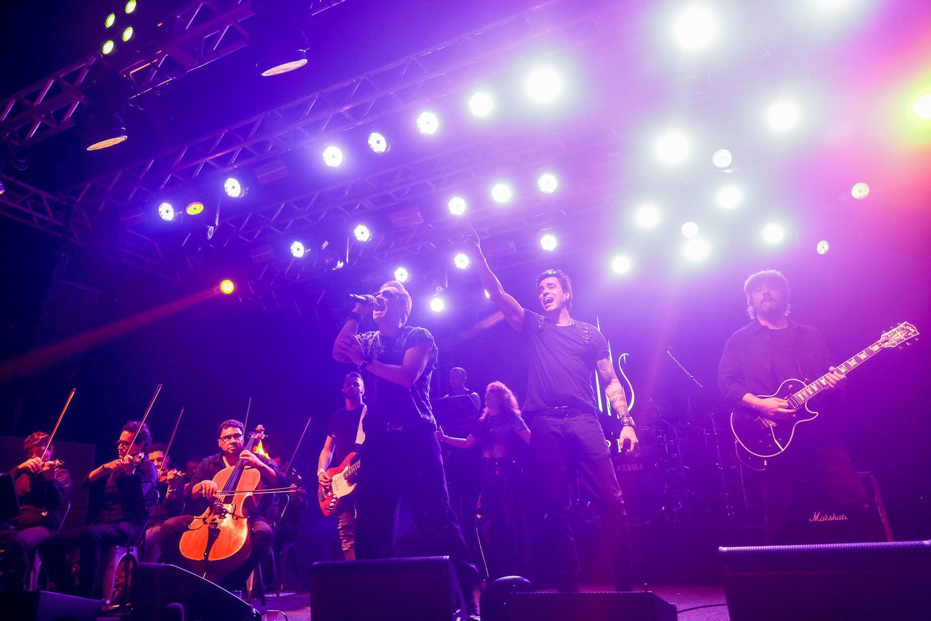 Uma banda se apresenta no palco, iluminada por refletores brilhantes. Os músicos tocam instrumentos, os vocalistas cantam.