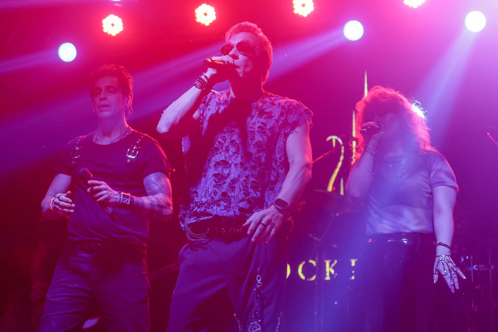 Três pessoas cantando no palco sob luzes de palco brilhantes.