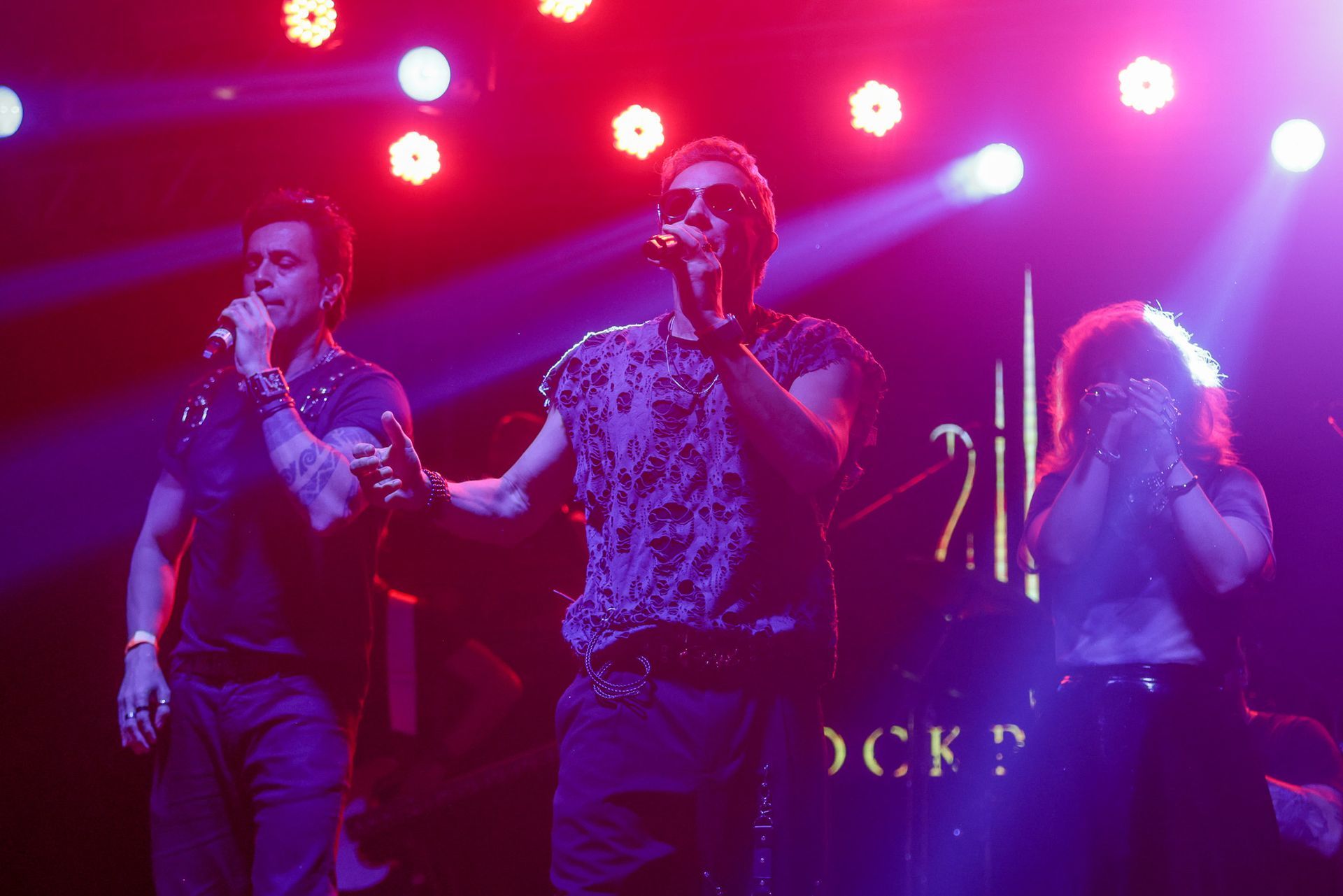Três pessoas no palco, cantando. Luzes de palco roxas e vermelhas as iluminam.