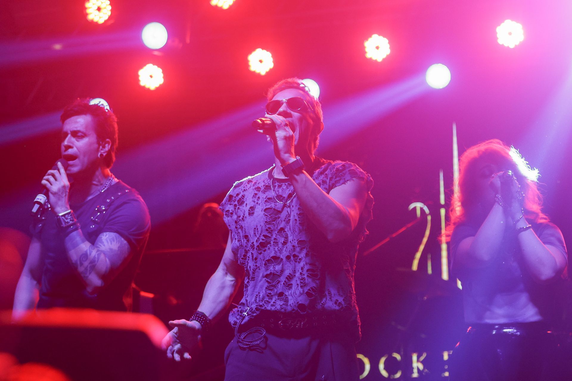 Três pessoas no palco, cantando. Luzes de palco roxas e vermelhas as iluminam.