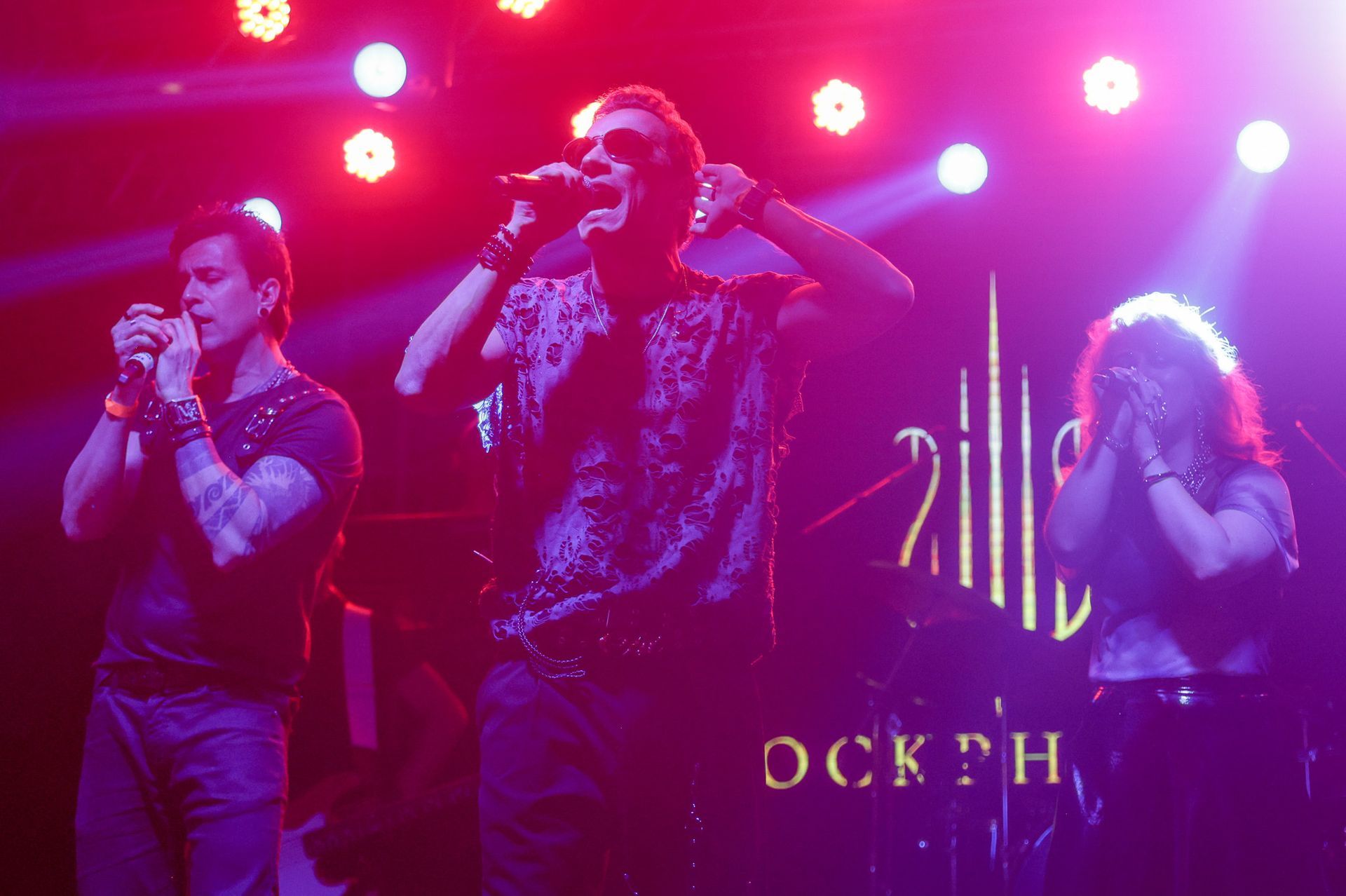 Três pessoas no palco, cantando. Luzes de palco roxas e vermelhas as iluminam.