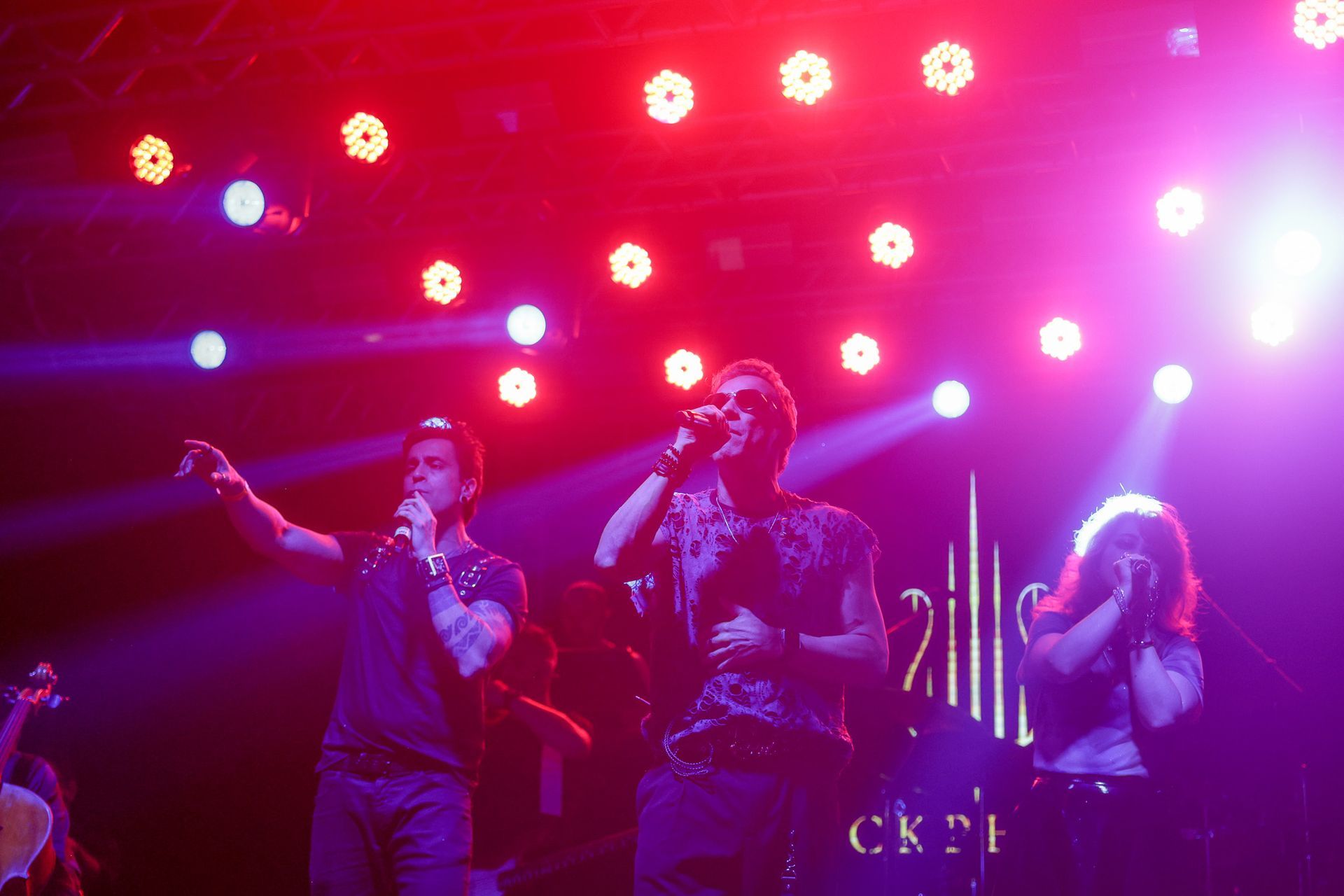 Três pessoas no palco, cantando. Luzes de palco roxas e vermelhas as iluminam.