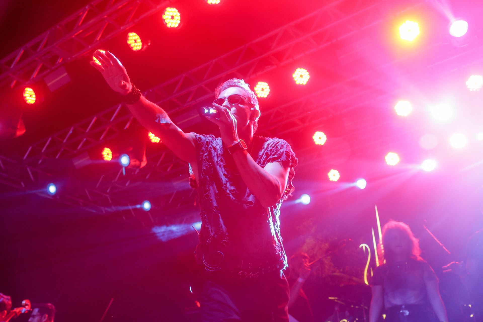 Cantor se apresentando no palco com o braço levantado, iluminada por luzes de palco vermelhas e brancas.