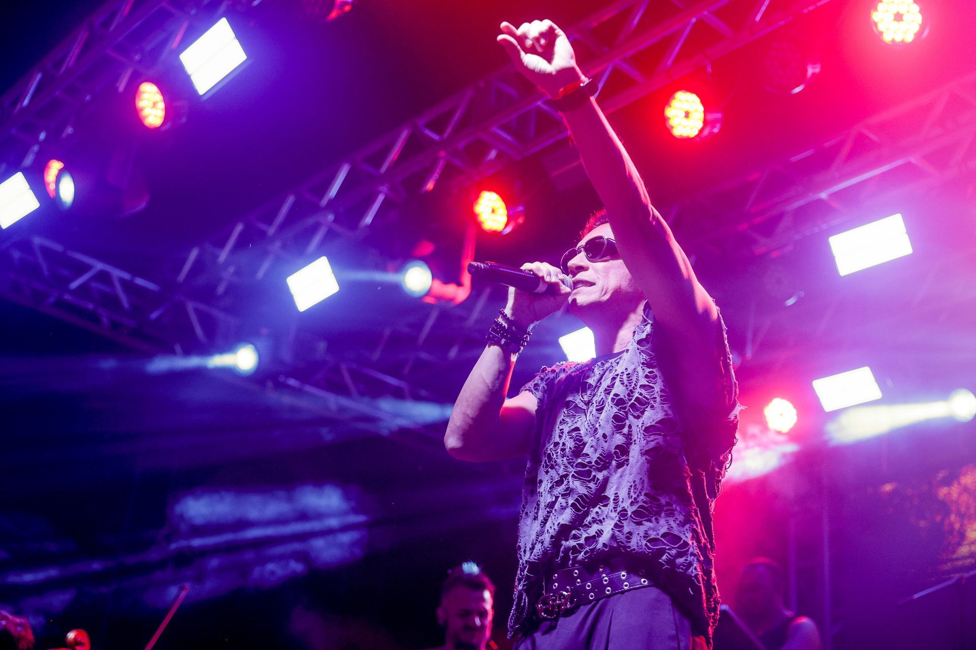 Homem cantando em um microfone no palco, com os braços erguidos, sob luzes de palco brilhantes.
