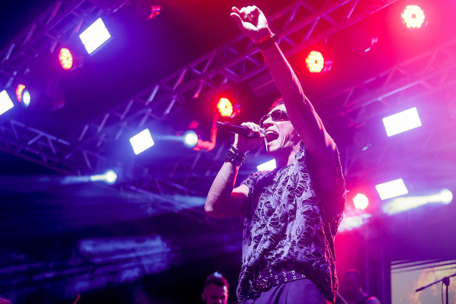 Um artista cantando em um microfone no palco, com o braço levantado. Luzes de palco roxas e vermelhas se acendem.