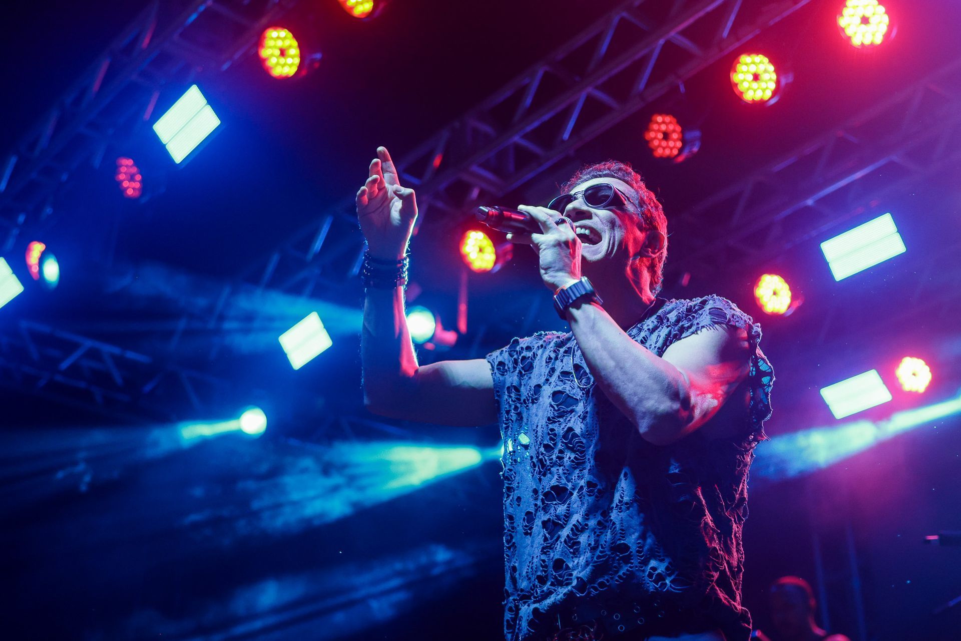 Cantor no palco, iluminada por luzes azuis e vermelhas, segurando um microfone, com o braço levantado