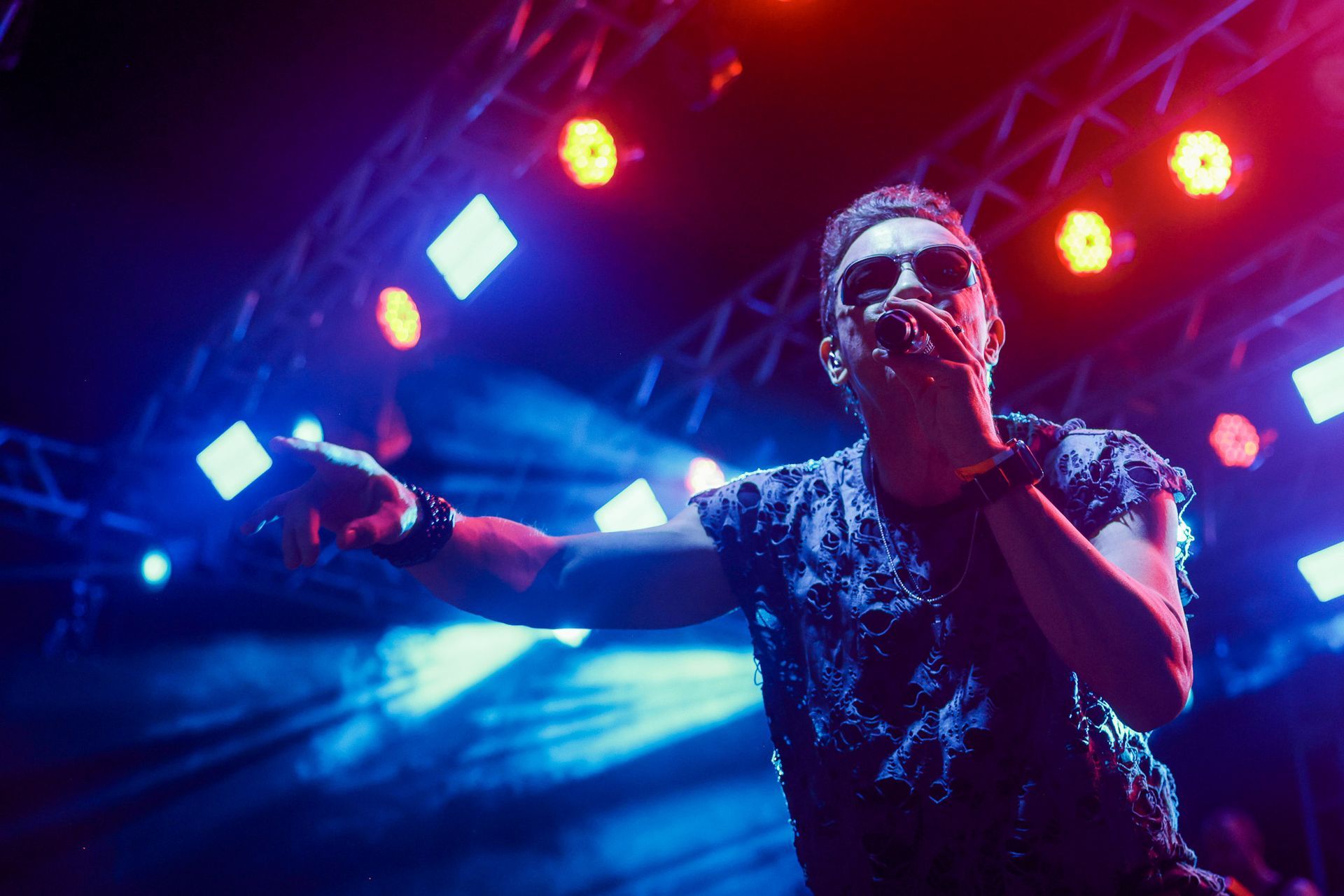 Cantor no palco, iluminada por luzes azuis e vermelhas, segurando um microfone, com o braço levantado