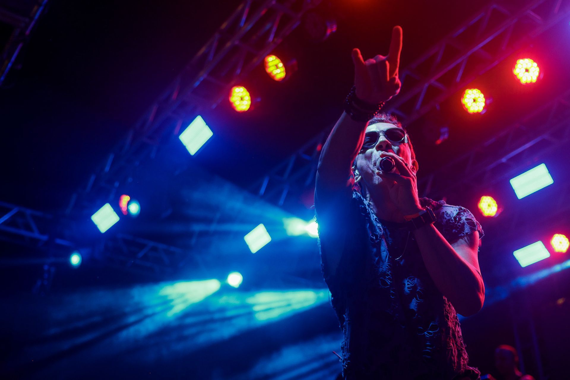 Cantor no palco, iluminada por luzes azuis e vermelhas, segurando um microfone, com o braço levantado