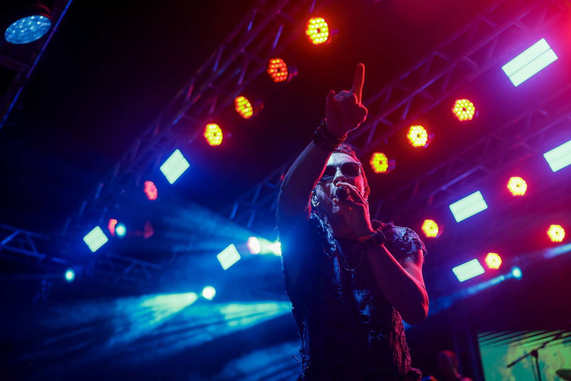 Cantor no palco, iluminada por luzes azuis e vermelhas, segurando um microfone, com o braço levantado