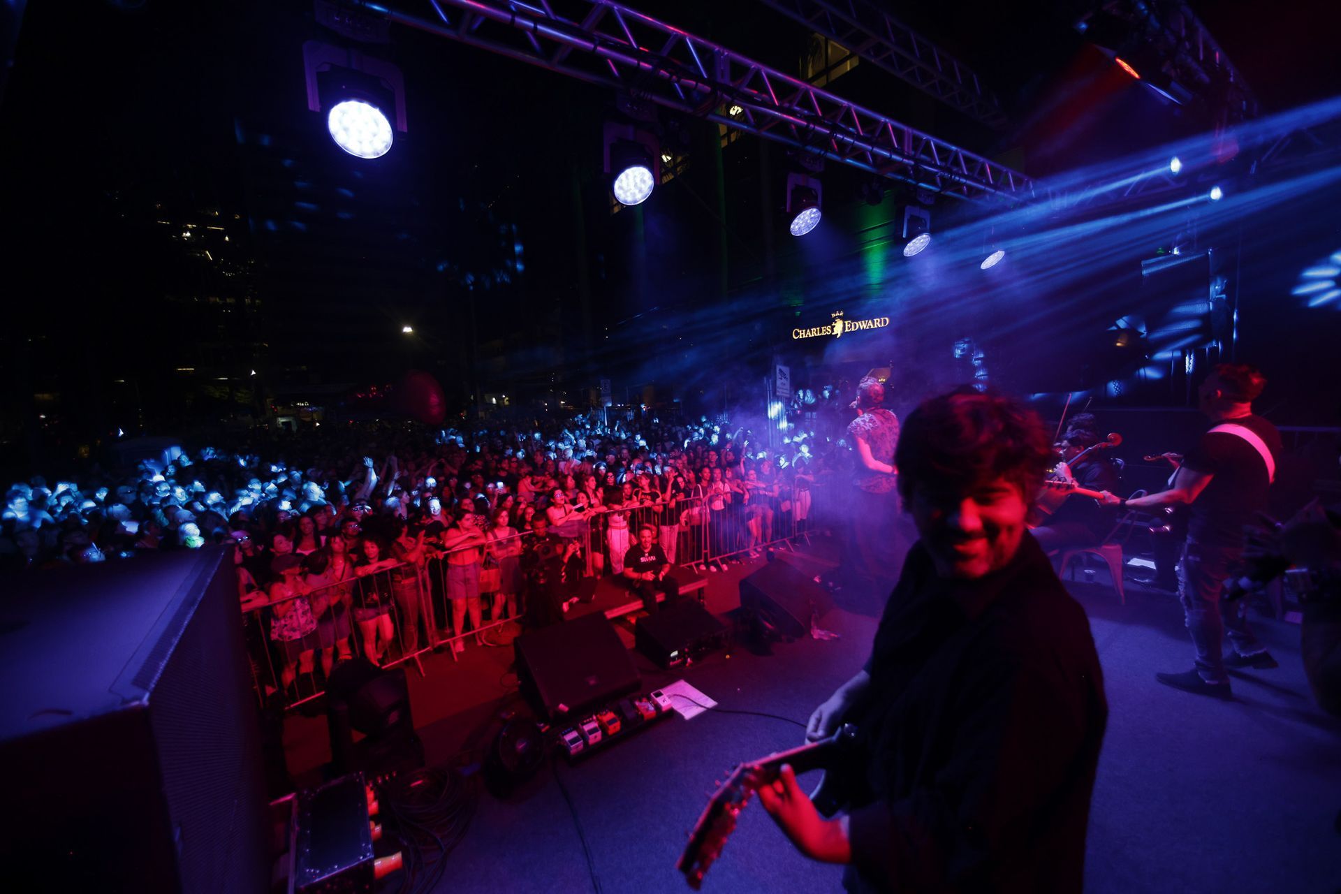 Banda se apresentando no palco durante um show. Público ao fundo, iluminado com luzes azuis e vermelhas.