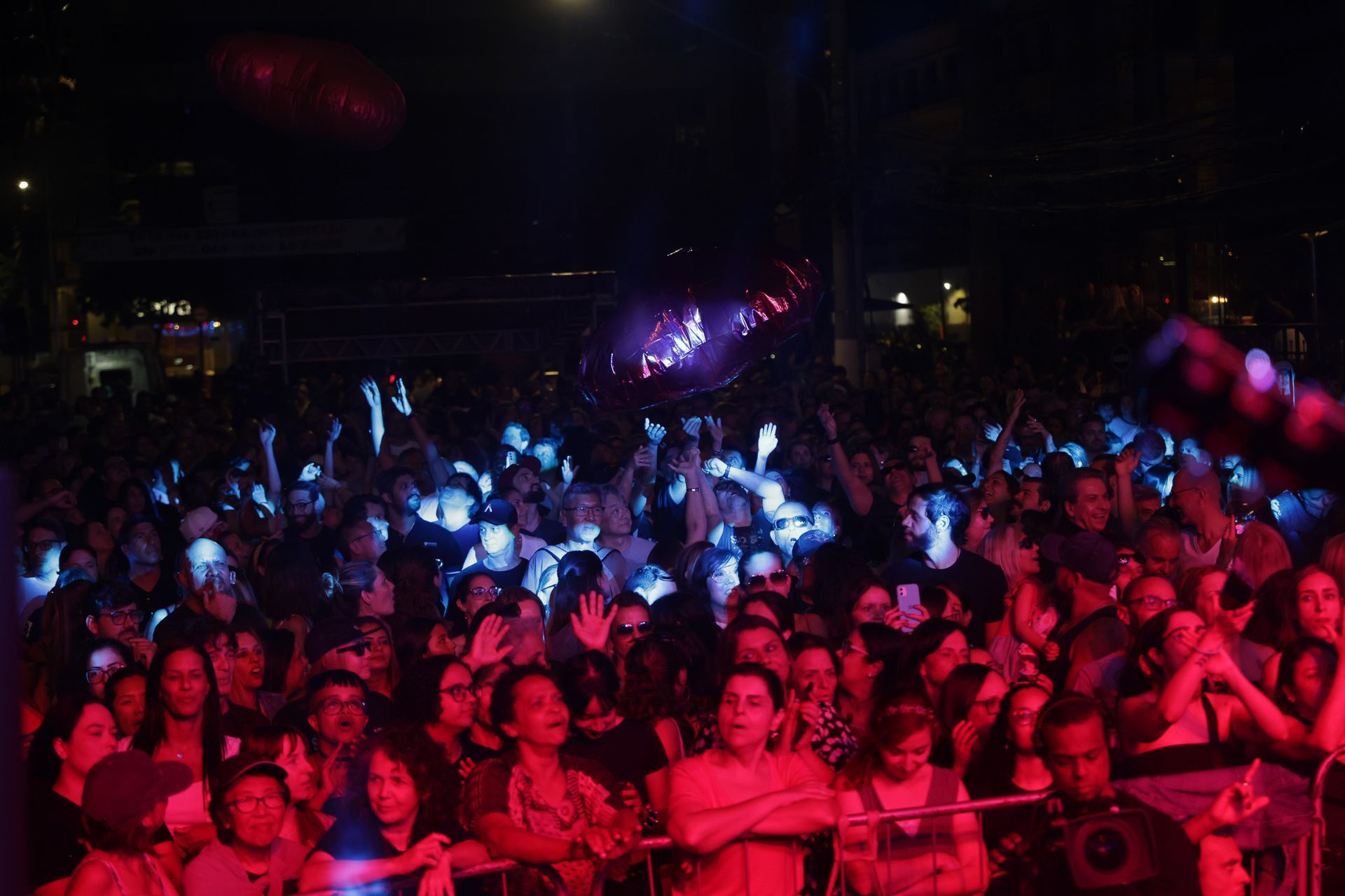 Público em um show com luzes azuis e vermelhas; mãos erguidas; ambiente noturno.