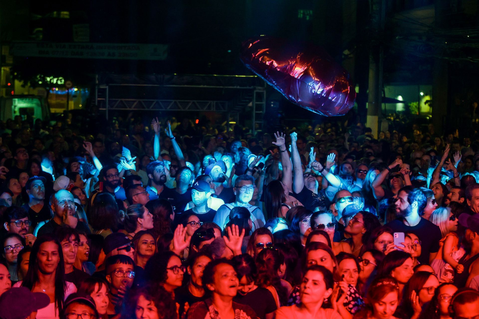 Público em um show ao ar livre, iluminado por luzes de palco azuis e vermelhas. As pessoas levantam os braços.