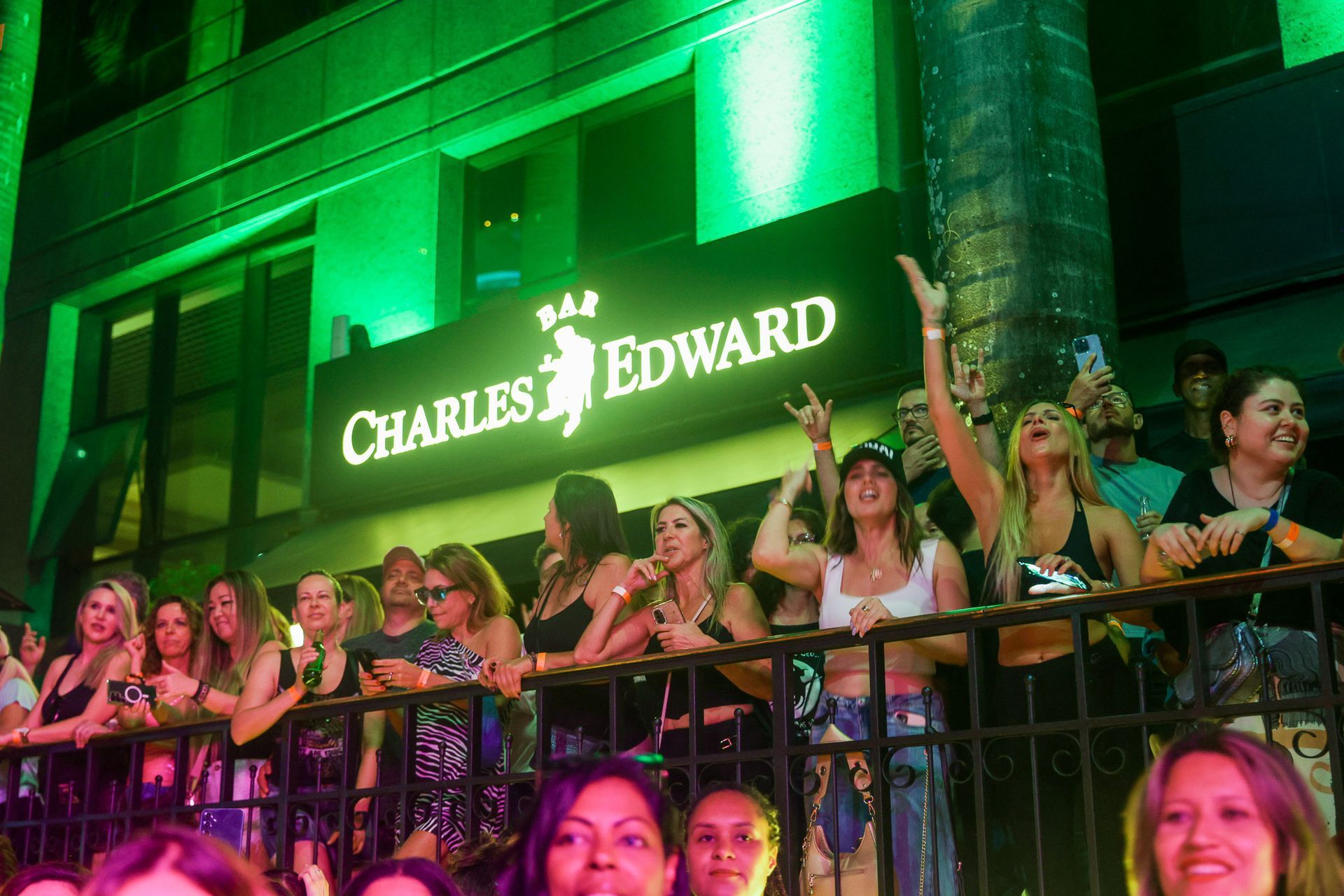 Multidão do lado de fora do bar Charles Edward, iluminado em verde. As pessoas sorriem e mostram seus celulares.