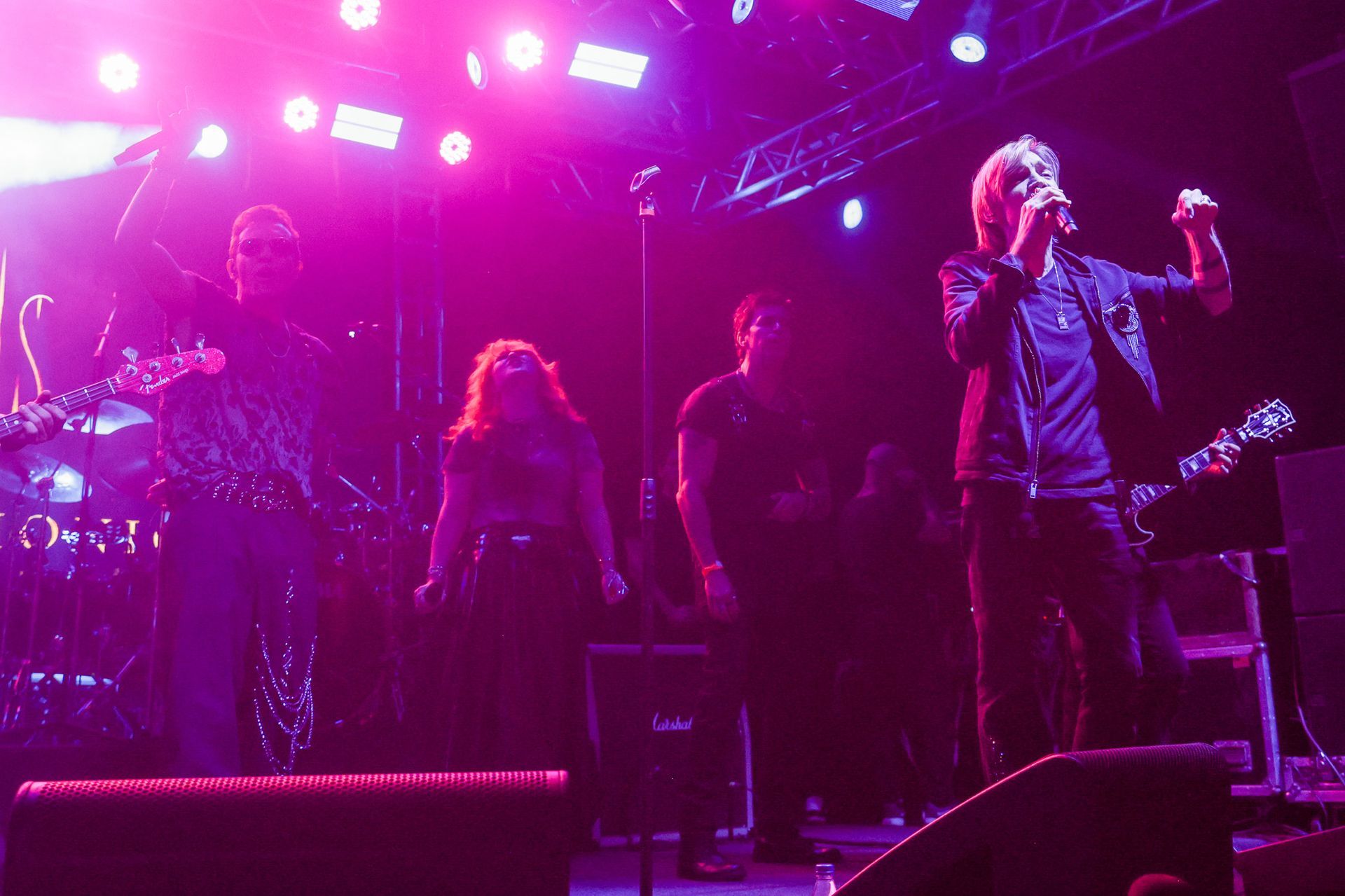 Banda se apresentando no palco sob luzes roxas. Vocalista com o braço levantado, outros tocando instrumentos.