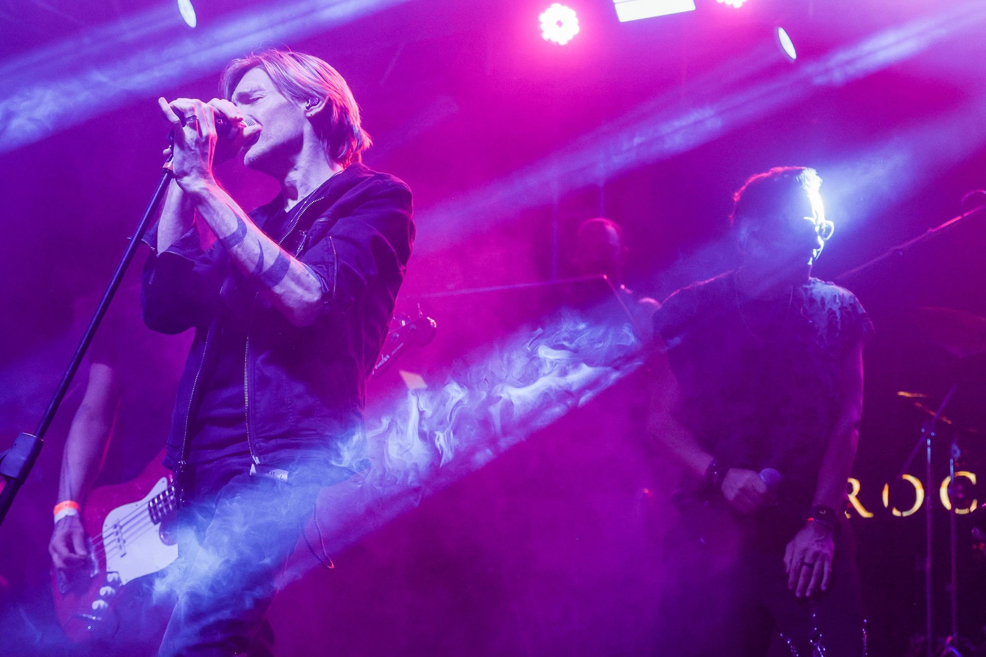 Cantor se apresentando no palco sob holofotes brilhantes, outro músico ao fundo. Luzes roxas e brancas.
