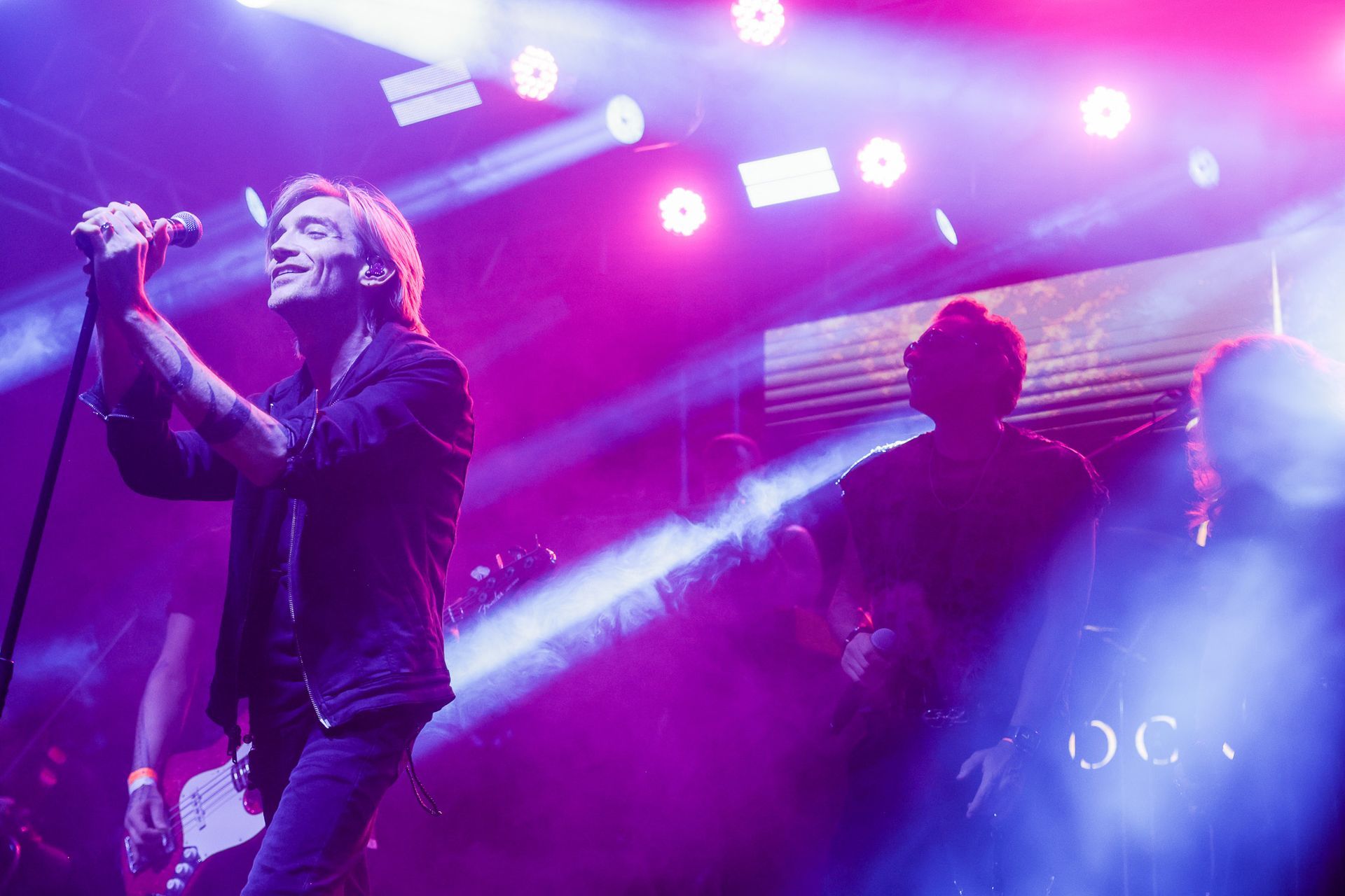 Cantor se apresentando no palco sob holofotes brilhantes, outro músico ao fundo. Luzes roxas e brancas.