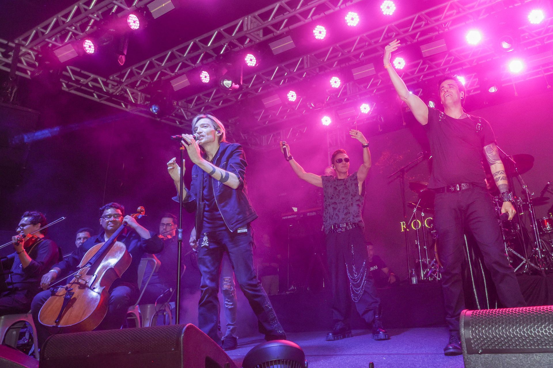A banda se apresenta no palco sob luzes rosas com orquestra. Dois integrantes da banda levantam os braços.
