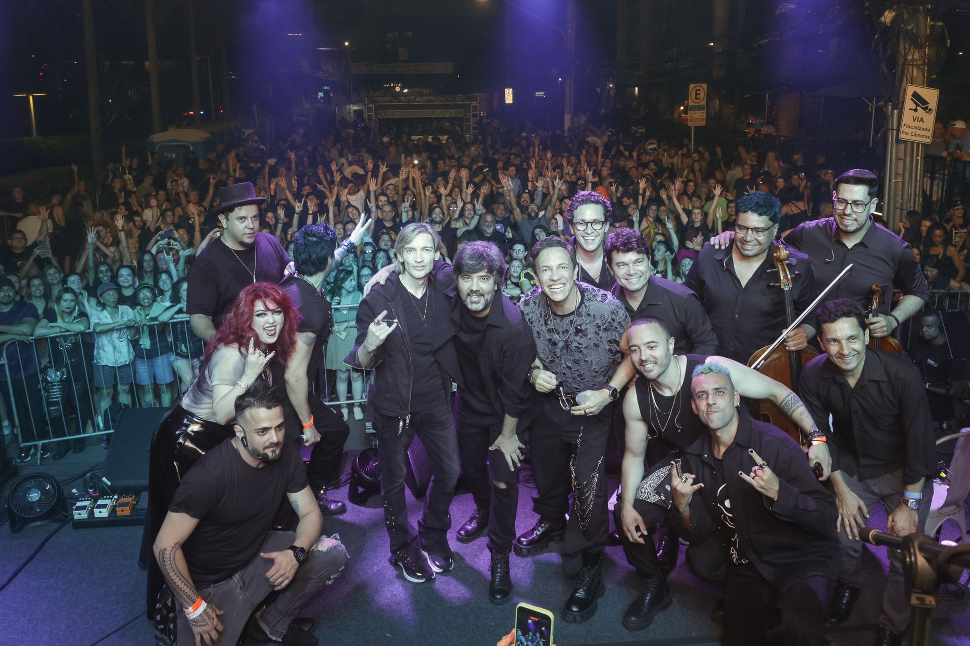 A banda posa para fotos com o público em um show ao ar livre; os músicos vestem roupas escuras e fazem gestos com as mãos.